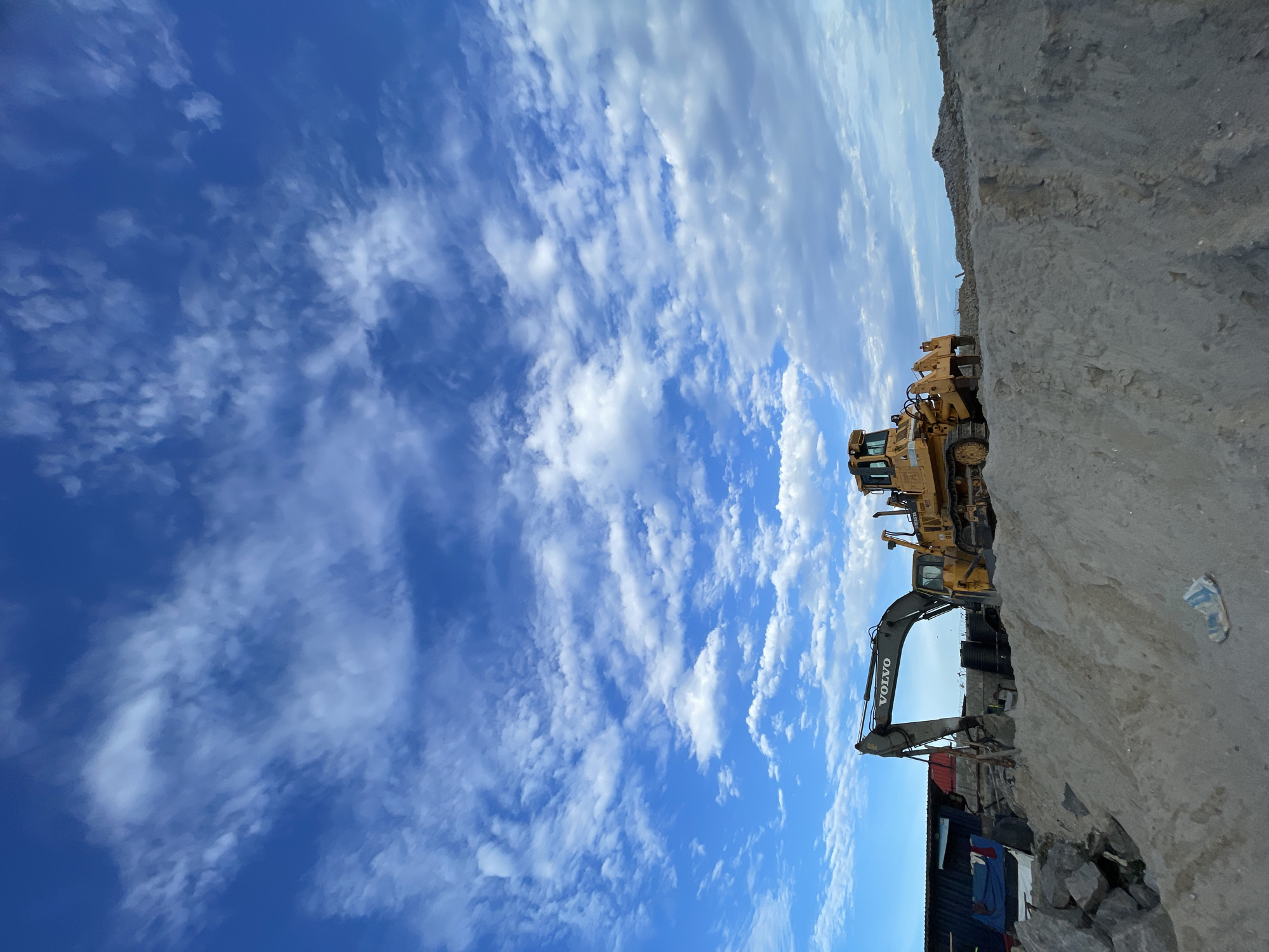The image depicts a construction or excavation site in Ilado, Nigeria. Dominating the foreground is a large earth mound. A Volvo excavator and a bulldozer are positioned on top of the mound. In the background, there is a dark, container-like structure. The sky is blue with scattered white clouds.