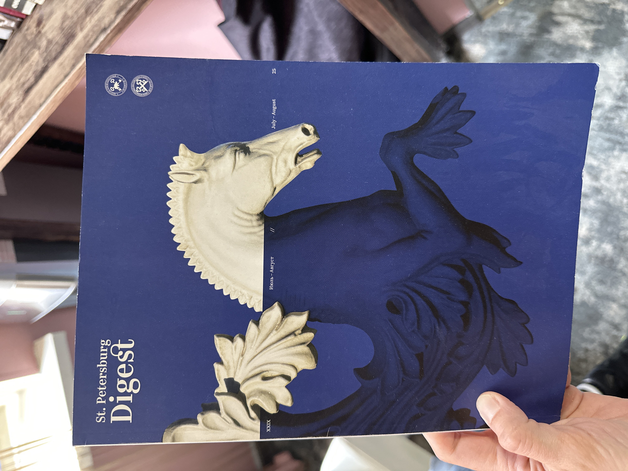 A close-up photograph depicts a copy of "St. Petersburg Digest" magazine held by a person. The cover features a stylized image of a horse figure split into light and dark halves against a dark blue background. Text on the cover includes "St. Petersburg Digest" written vertically on the left side, the publication months "July-August," the number "25" at the top right, the name "Hans-Auyer," and the number "XXIX" at the bottom left. Two circular logos are located at the top left corner of the cover. The background of the image shows a wooden surface, a dark fabric garment, and a hand holding the magazine. The lighting is diffused.