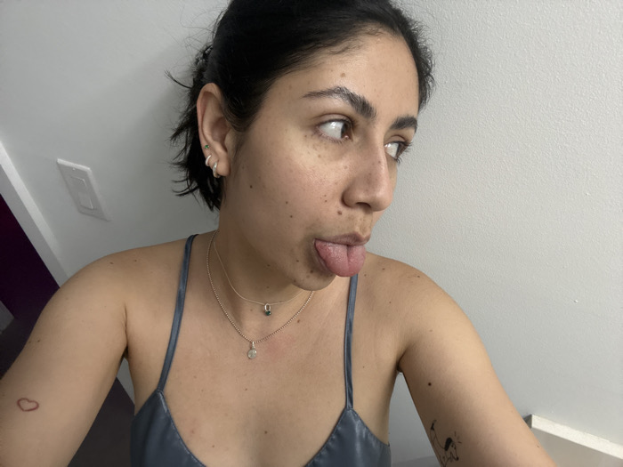 A woman poses playfully with her tongue out and eyes glancing to the side. She wears a camisole, and two delicate necklaces. Her dark hair is pulled back. She has a light complexion and freckles. A small heart tattoo is visible on her arm, along with another cartoonish tattoo. She has multiple earrings in her left ear. The background consists of a light-colored wall and a portion of a dark-colored area on the left. A light switch is also visible on the wall.