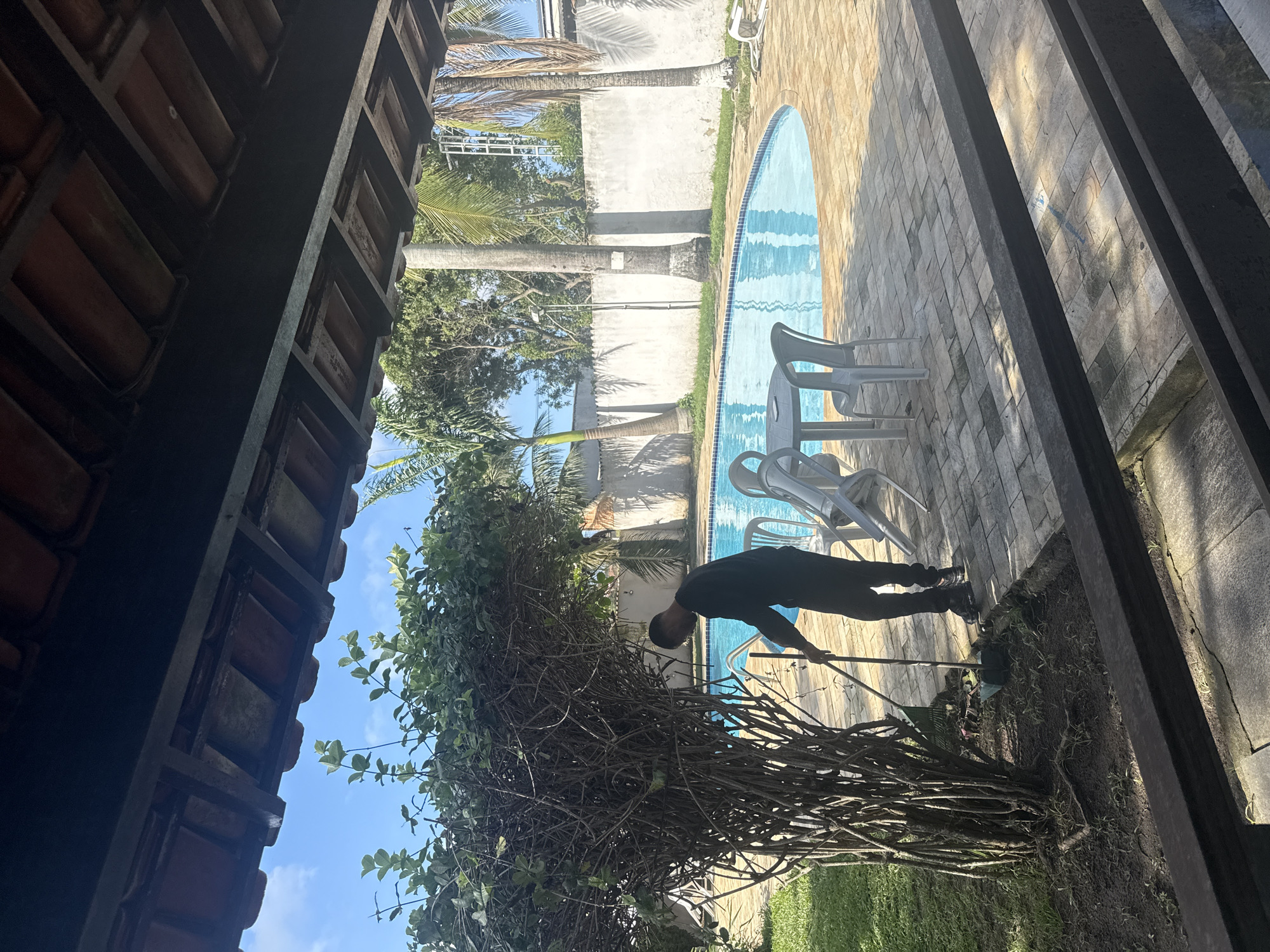 A man is cleaning the area around a large bush next to a swimming pool. He is wearing a black long-sleeved shirt and black pants. He is using a broom and dustpan to gather debris from the ground, which appears to be soil and dry leaves.
The swimming pool is oval-shaped and filled with clear blue water. It is surrounded by a brick patio. There are several white plastic chairs and a round table with chairs on the patio. Palm trees and other greenery are visible in the background, as is a white wall. The scene appears to be located at a residence or a resort in Cabo Frio, Brazil. The time of day is likely daytime, given the bright sunlight and shadows.