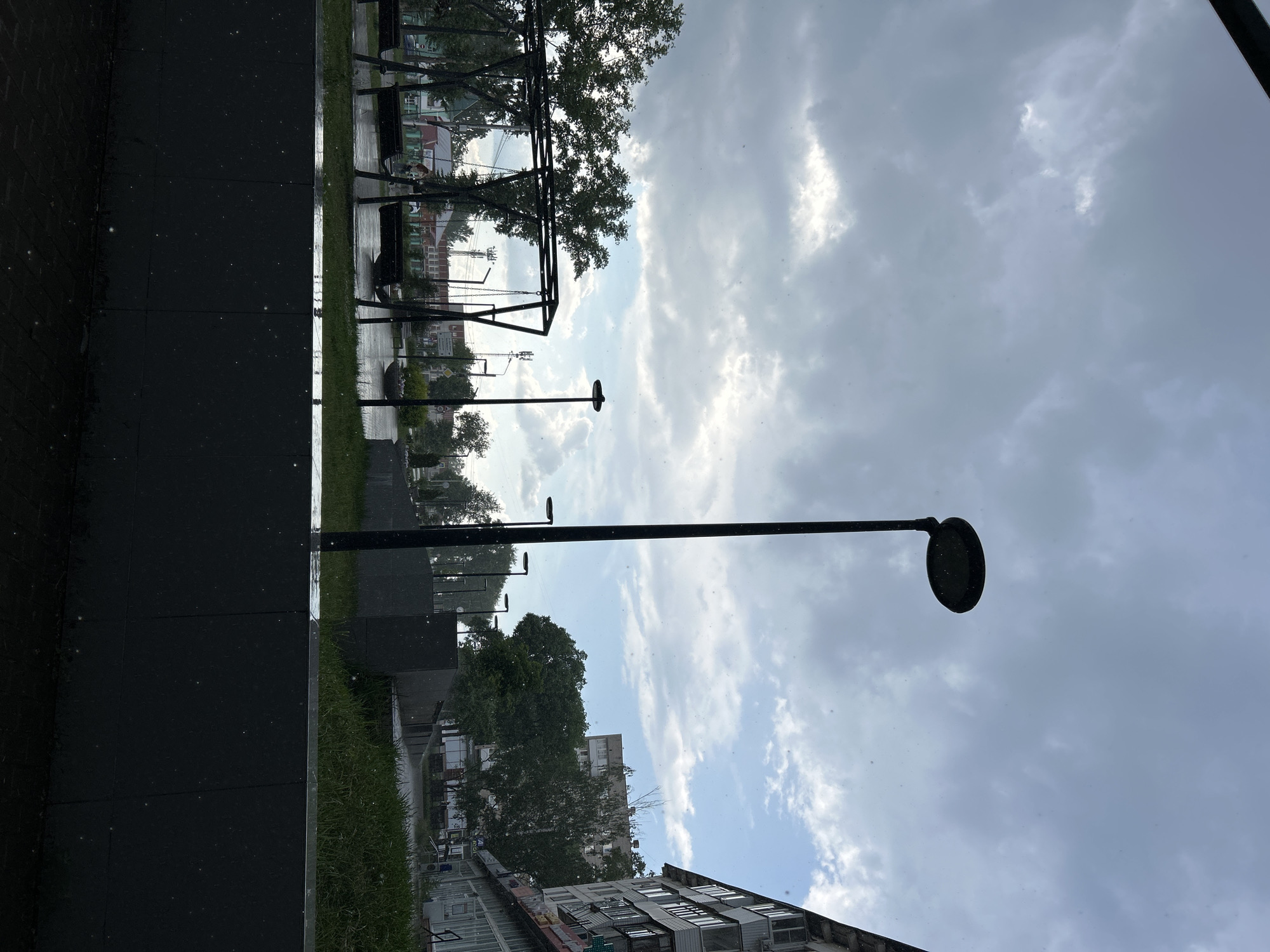 The image captures a street scene in Chaikovsky, Russia, on a cloudy day with precipitation visible as small white dots. In the foreground, a dark grey, textured wall runs horizontally across the frame. Beyond this wall, a children's playground with swing sets is visible on the left, featuring black metal structures and empty swings. To the right of the playground, a tall, black lamppost stands prominently.
Further in the background, several more lampposts are sparsely distributed. Green trees line the area behind the playground. Buildings of varying heights and architectural styles are visible, including multi-story apartment buildings on the right side of the frame and what appear to be smaller structures further in the distance. The sky is overcast with a mix of grey and lighter clouds, suggesting a damp atmosphere. The ground in the foreground appears to be paved with dark interlocking bricks, wet from the precipitation. The overall scene depicts an urban public space during inclement weather.