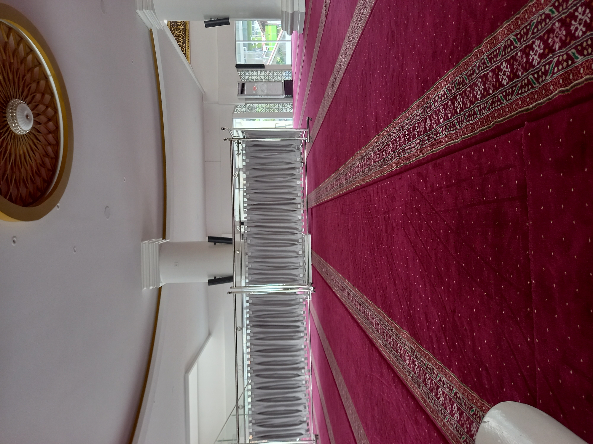 An interior photograph shows the interior of a mosque with red carpeted floors. The carpet has a decorative border in gold and red. In the background, a metallic partition with grey curtains separates an area of the mosque. Large white pillars with ornate gold trim and a decorative ceiling are visible. Several speakers are mounted on the pillars. The scene appears to be in Samarinda, Indonesia.