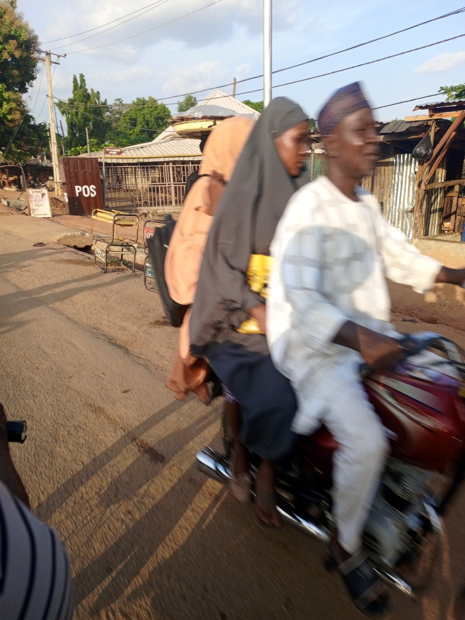 Three people are riding a red motorcycle on a paved road in Keffi, Nigeria. The driver is wearing a white and blue striped shirt and a blue cap. The middle passenger wears a black hijab, while the third passenger is wearing a peach-colored head covering. A "POS" sign is visible on the left side of the road, along with other buildings and utility poles.