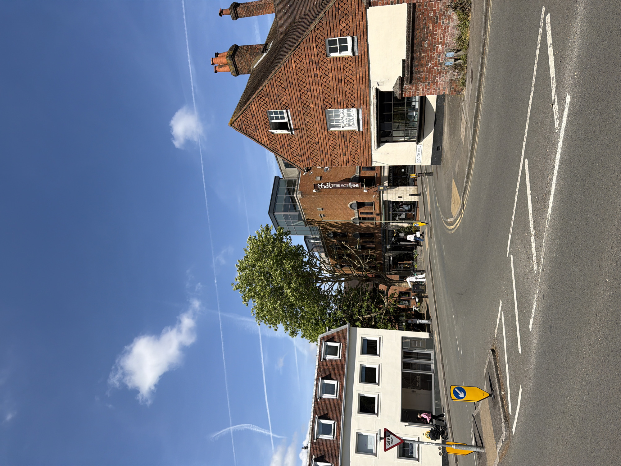 The image depicts a street scene in Guildford, United Kingdom. The view is upwards toward buildings on the side of the road.
Buildings:
- A red brick building with a steeply pitched tiled roof and chimneys, including a white sign that says "SOUTH HILL".
- A modern brick and glass building housing "Thai Terrace" restaurant.
- A white building with multiple rectangular windows and a brown tile roof.
Street:
- An asphalt road marked with white directional arrows and yellow road markings, including a traffic sign that directs traffic to the left.
Other:
- A large green tree partially obscures the view of some buildings.
- A blue sky is visible with several contrails.
- A stroller is on the sidewalk.