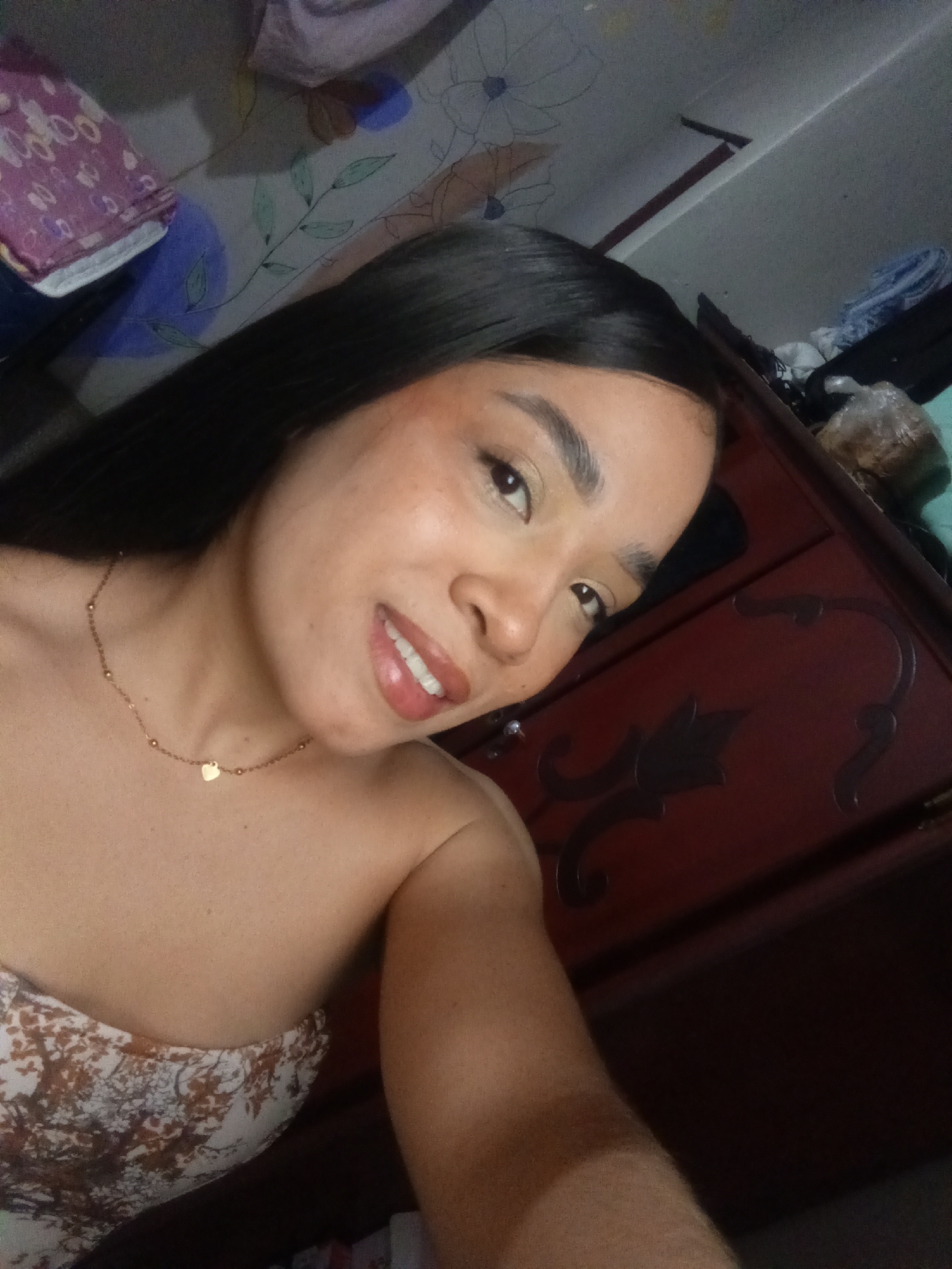 A woman is taking a selfie indoors in what appears to be a bedroom. She has long, dark hair and is wearing a strapless patterned top and a delicate gold necklace with a heart pendant. She is smiling slightly. Behind her is a dark wood dresser and some cluttered items on top, including what looks like folded clothing. The ceiling and part of the wall are visible in the upper background, including a floral pattern painted on the ceiling.
The location in Valle de La Pascua, Venezuela cannot be determined.