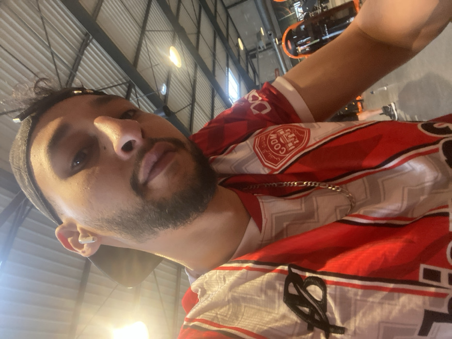 A mid-shot selfie depicts a male individual within an indoor fitness center, specifically identified as a Basic-Fit gym located at 10 Avenue du Général de Gaulle in Hœnheim, France. The subject, a male with a beard and dark hair, wears a black baseball cap reversed and a white wireless earbud in his right ear. He is clad in a red and white striped sports jersey, worn over a white t-shirt, featuring a black "AB" logo on the left chest and a circular emblem on the right sleeve with Arabic text and a mosque motif, along with "COD M". A silver chain necklace is visible around his neck. The background reveals a high industrial ceiling with exposed metallic beams, ductwork, and various lighting fixtures, including both recessed and pendant lights. Exercise equipment with prominent orange accents, including a piece of "MATRIX" branded machinery, is visible to the right of the frame. The floor is a light, uniform color. Faint shapes of other individuals and equipment are discernible in the deeper background.