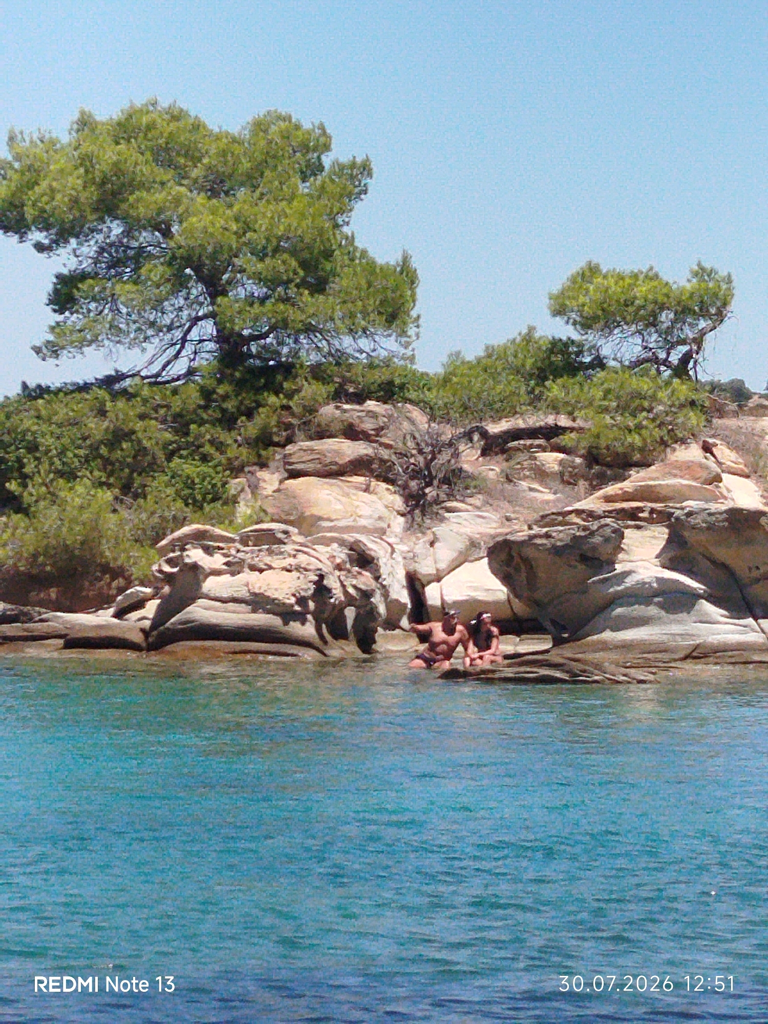 The image depicts a coastal scene in Sithonia Municipal Unit, Greece, characterized by turquoise waters lapping against rocky shores. Several individuals are visible, seated on the rocks at the edge of the water. One individual, appearing to be a male, is partially submerged in the water and is looking towards another individual, seemingly a female, who is seated on the rocks. The landscape features lush green trees and vegetation on the rocky terrain, with a clear blue sky overhead. The presence of the date and time stamp "30.07.2026 12:51" and "REDMI Note 13" suggests this is a photograph taken with a mobile device.
