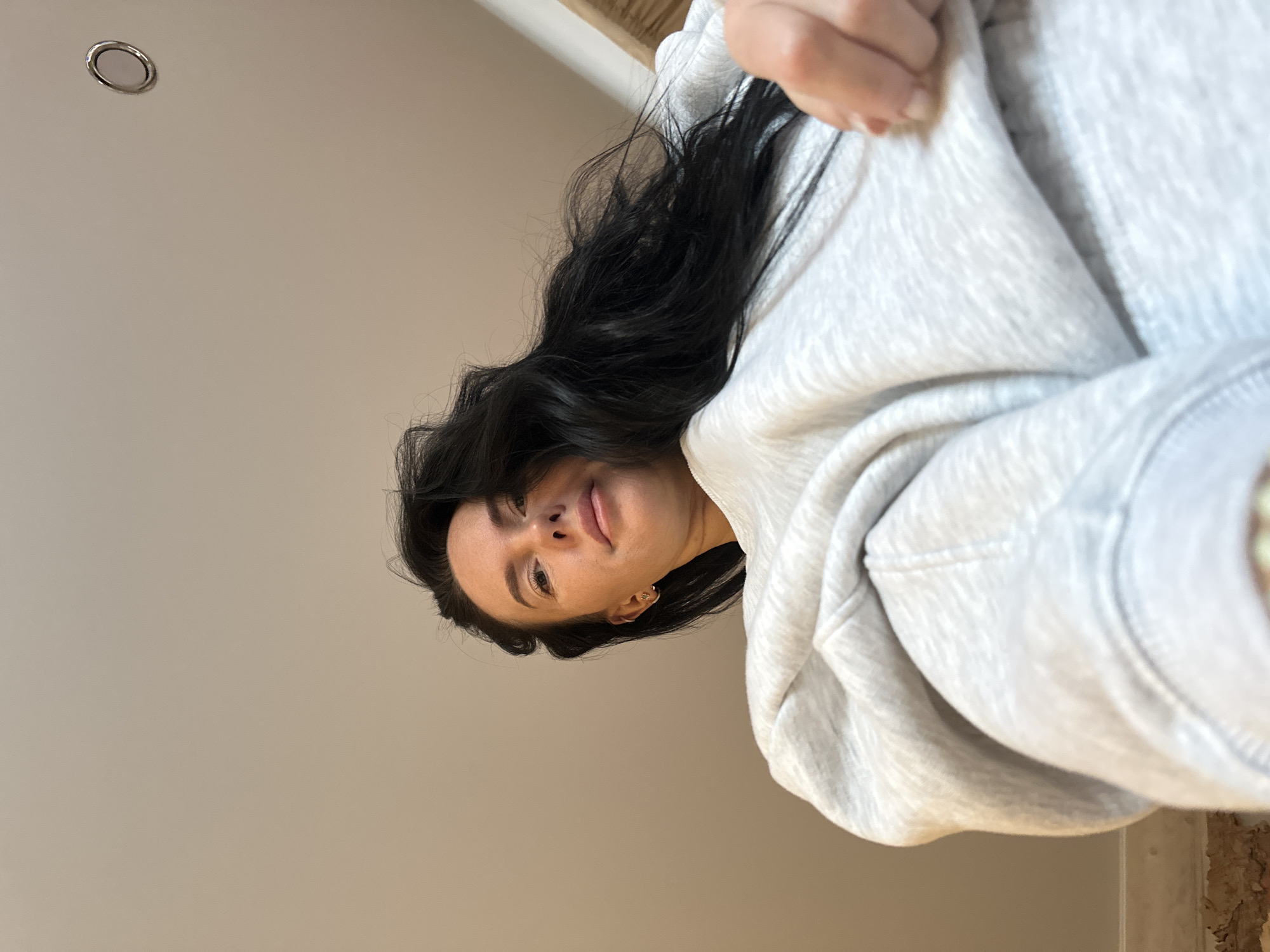 A fair-skinned woman with dark hair, wearing a light gray hoodie, is looking down at the camera. She is indoors, standing below a ceiling with a recessed light fixture. The background wall is a pale beige color. The location is unknown and cannot be determined from this image.