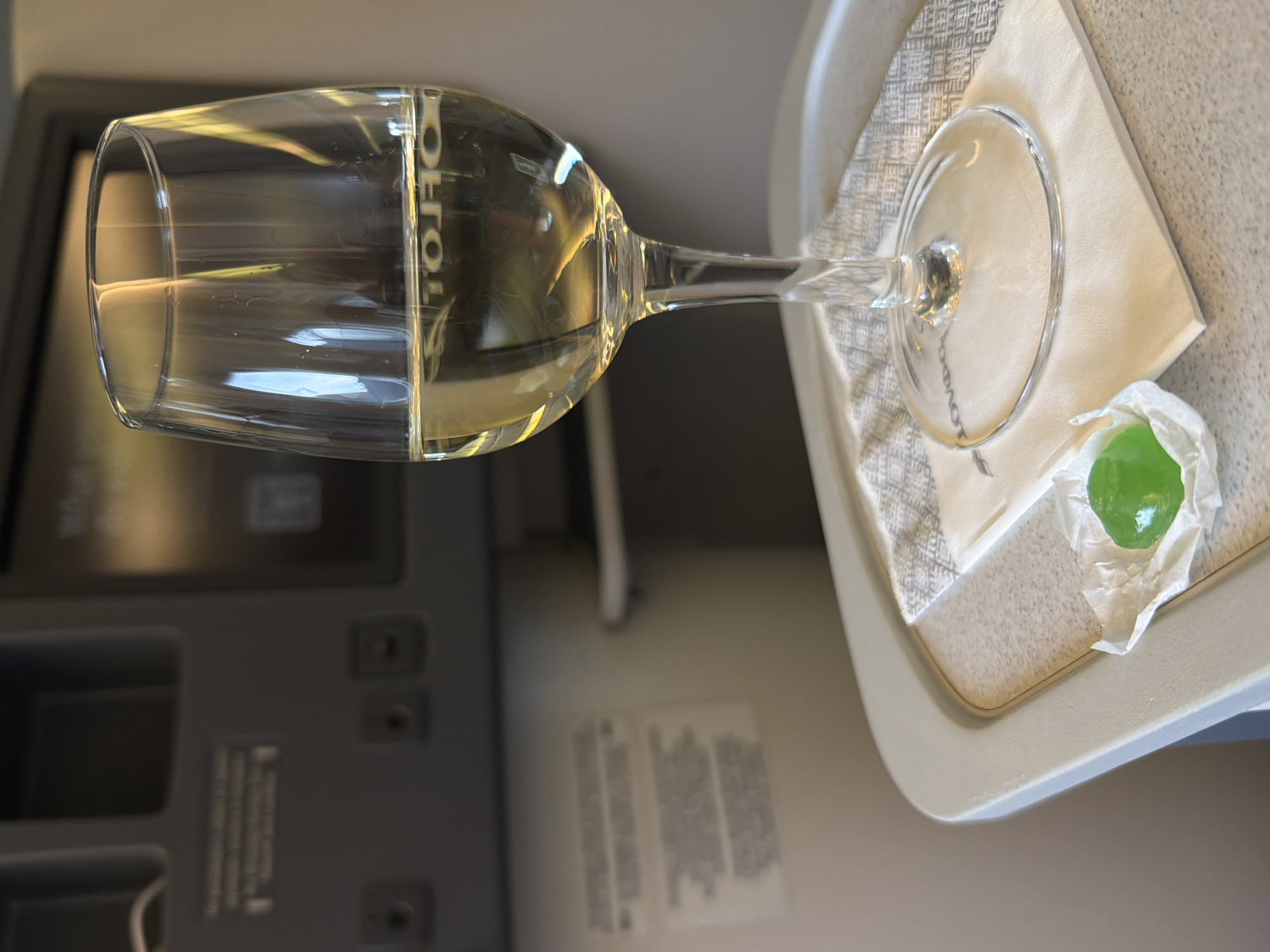 The image shows a close-up of a wine glass filled with a light-colored liquid, placed on a tray in what appears to be an airplane cabin. A small green candy is unwrapped next to the glass. The tray also holds a white napkin with a patterned border. In the background, a seatback screen is visible, along with some control panels. The lighting suggests an indoor environment, possibly during daylight hours. The context of Yelizovo, Russia, does not appear to be reflected in any visual elements of the image.