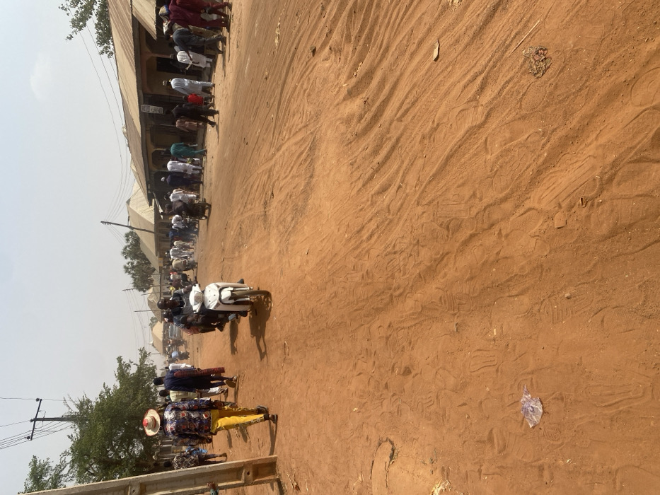 An unpaved, reddish-brown dirt street in Minna, Nigeria, is depicted under bright, hazy daylight. The ground is marked with numerous tire tracks and footprints. On the left, a utility pole with power lines stands beside sparse green trees. Along the right side of the street, a row of single-story buildings with corrugated metal roofs and plastered walls extends into the background. Approximately 30-50 individuals are present on the street. In the foreground on the left, a male subject walks away from the camera, wearing a straw hat, a multi-patterned long-sleeved shirt, and bright yellow trousers. In the center-left, a male subject operates a white scooter-style motorcycle, riding towards the viewer. Another motorcycle with a rider is visible further back on the right. Individuals are seen walking, standing, and riding motorcycles, indicating general street activity or a gathering. Sunlight casts distinct shadows.