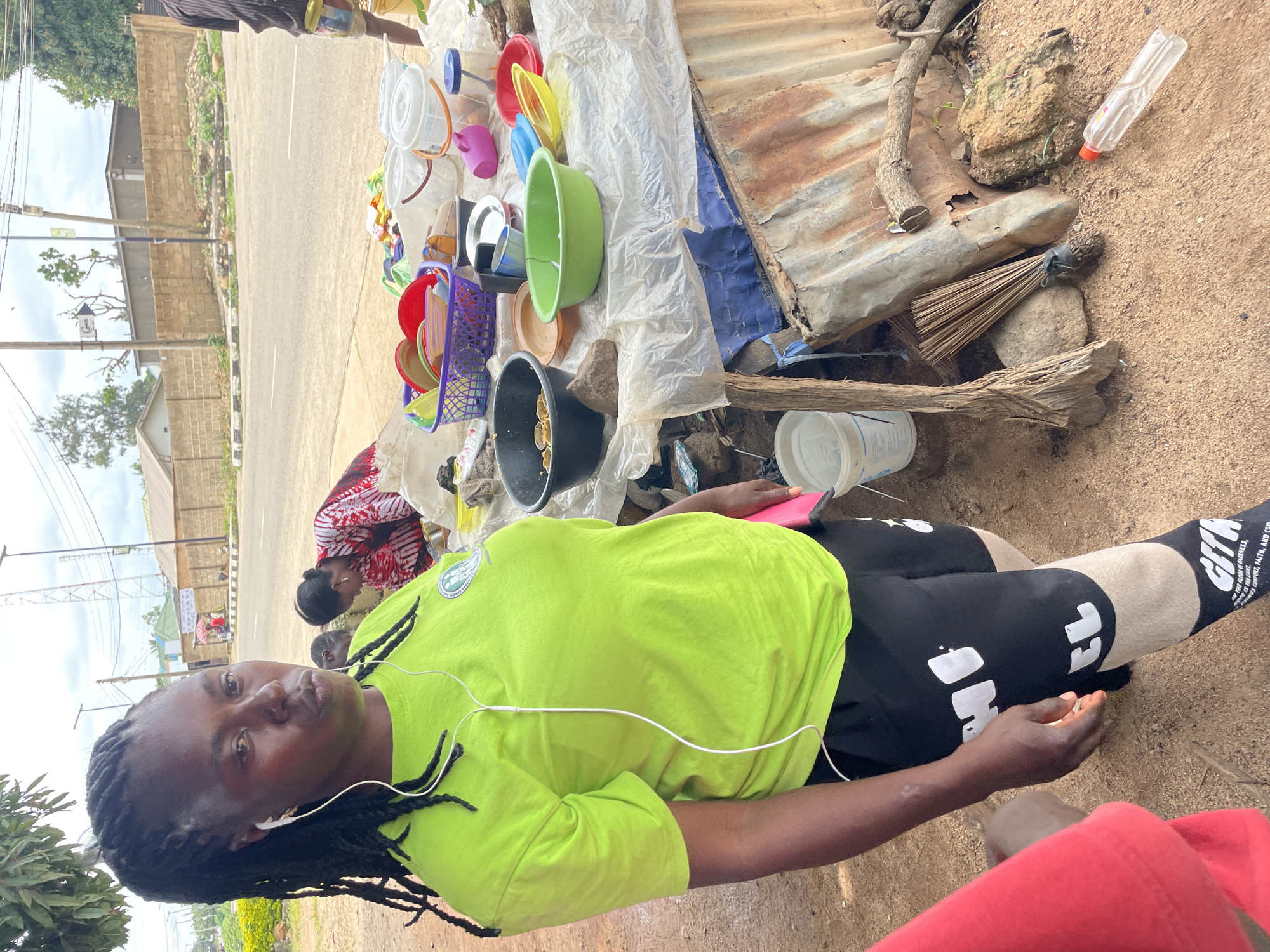 A medium shot shows a woman wearing a bright green t-shirt and black shorts standing on a dirt road in Jos, Nigeria. She has braided hair and is wearing white earbuds. Behind her, a table displays various household items, including plastic bowls, plates, and buckets. A few people are visible in the background, including a woman in a red and white patterned dress. The scene appears to be a roadside market. The overall environment suggests a warm, possibly humid climate.