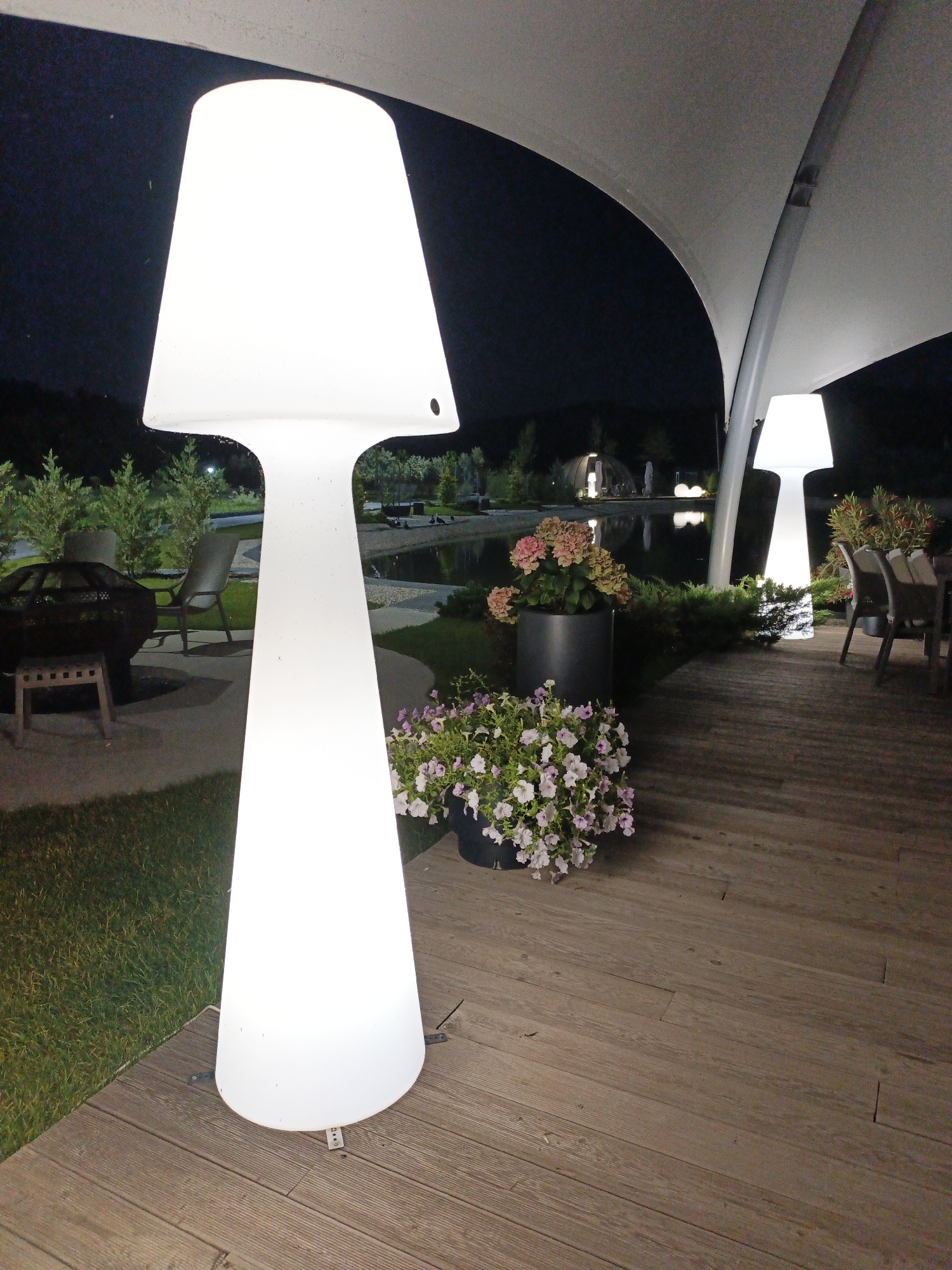 This image depicts an outdoor seating area at night, likely a patio or deck. The primary subjects are two large, illuminated, white mushroom-shaped lamps, one in the foreground and another further back. In the foreground, a cluster of pink and white petunias in a dark pot sits on the wooden deck, to the left of the large lamp. Behind the lamp, a dark metal fire pit with a wooden stool in front of it is visible on a paved area. Beyond this, a body of water, possibly a pond or lake, reflects the night sky and distant lights.
In the background, more lighting can be seen, including another illuminated lamp near a set of outdoor dining chairs and tables. The overall scene is illuminated by the artificial lights, suggesting it is nighttime. The sky is dark, with no visible stars, and the weather appears calm. The presence of a pond, fire pit, and seating suggests a relaxed, possibly resort or private property setting. There are no people visible in the image. The location is identified as Pshada, Russia. No text is visible in the image.