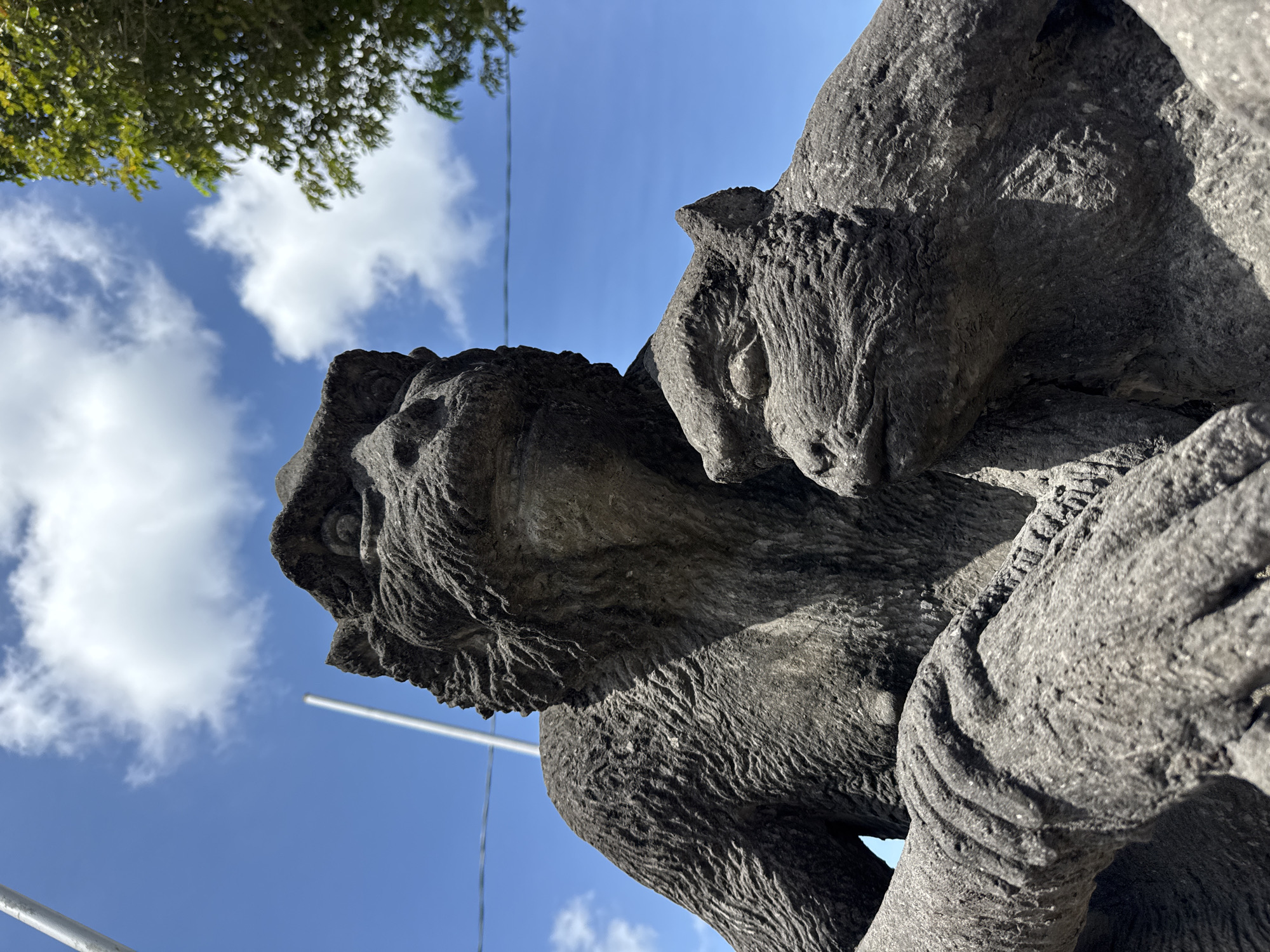 A low-angle shot shows a weathered stone statue of a man embracing a smaller animal, possibly a monkey or similar creature. The statue's texture is rough and heavily eroded. The sky is visible in the background, with patches of blue and scattered clouds. A sliver of greenery from nearby trees is seen at the top-left corner. The presence of poles in the background suggests the statue is in a public place. Based on the prompt, the location is likely Ubud District, Indonesia.