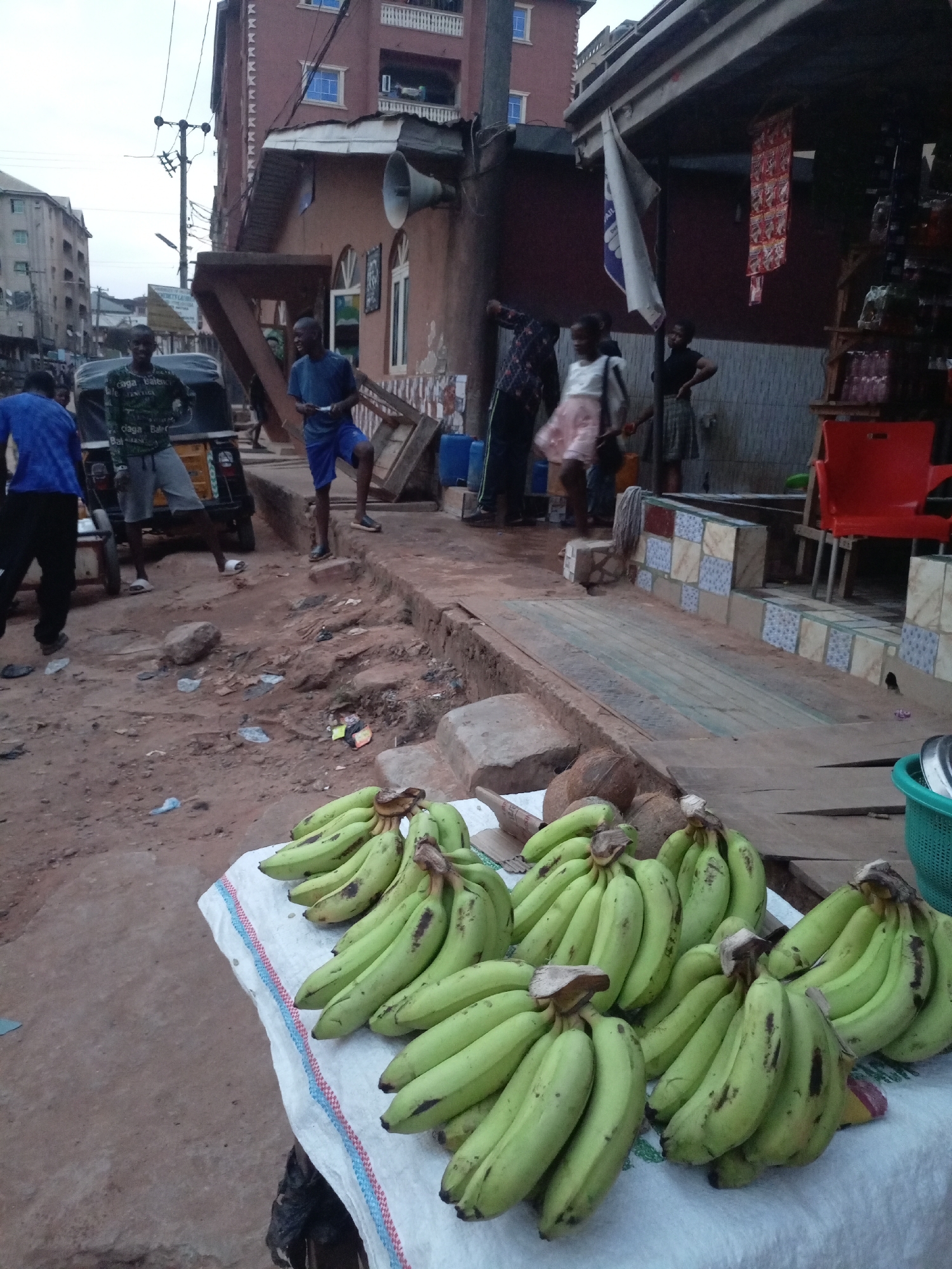 The image depicts a street scene in Onitsha, Nigeria, focused on a display of green plantains. Several bunches of plantains rest on a white surface, likely a table or cart. In the background, people are gathered near a building with a covered storefront. A motorized tricycle, known as a "keke," is parked nearby. The street surface appears uneven and partially paved. Overhead power lines are visible, contributing to the urban setting. A loudspeaker is attached to a building, and a flag hangs from the storefront. The overall environment suggests a bustling market or commercial area.