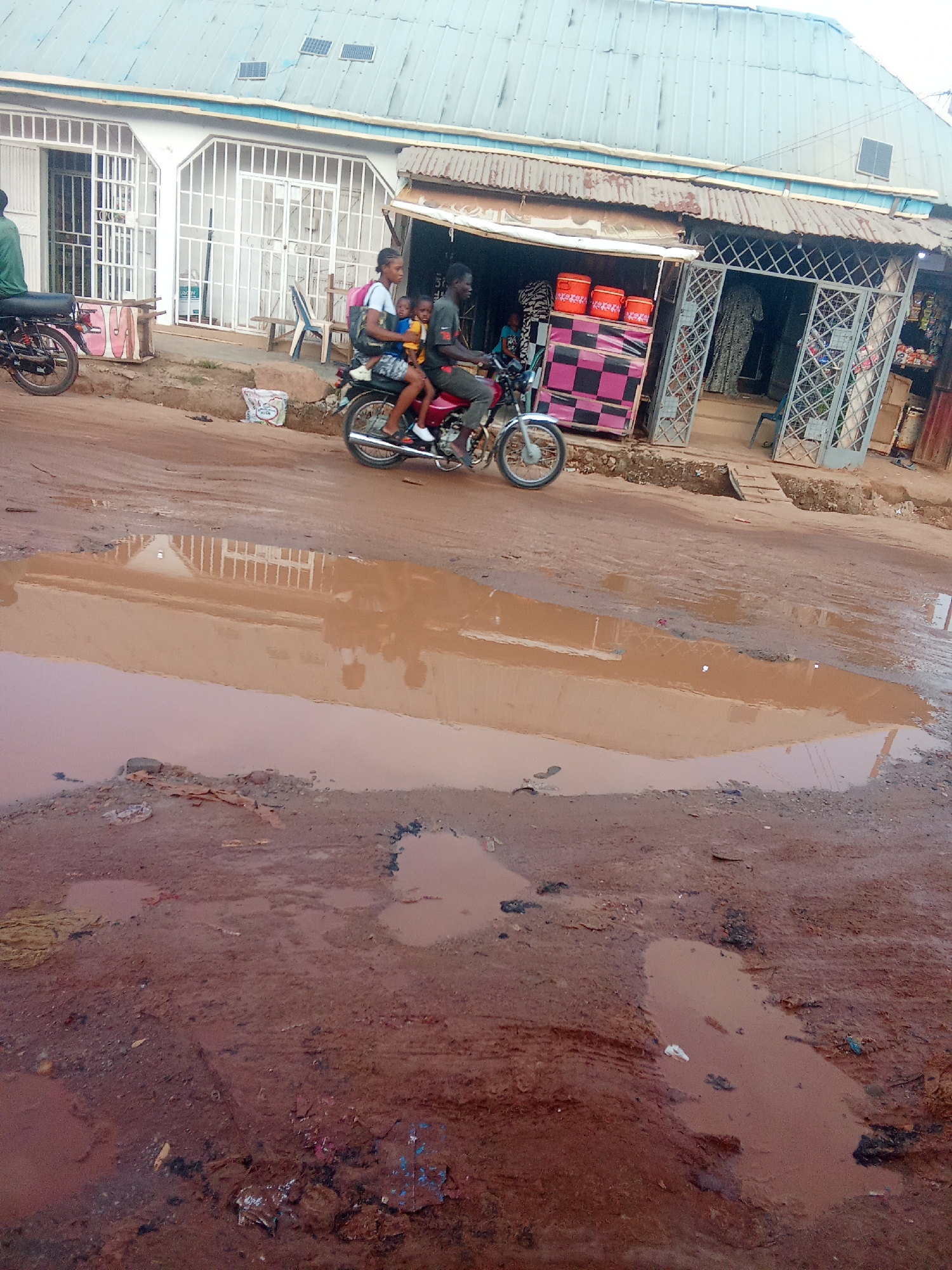 In Kagini, Nigeria, a family of three rides a red motorcycle through a muddy street. The motorcycle is driven by an adult male, with an adult female seated behind him and a young child between them. The motorcycle has a silver exhaust pipe and black tires. The street is dirt and filled with large puddles of brown water, indicating recent rainfall. A white plastic bag is visible on the side of the road. In the background, a building with a corrugated metal roof has an open storefront. The storefront displays orange coolers and a checkered display stand. A second motorcycle is parked to the left of the building. The sky is overcast.
