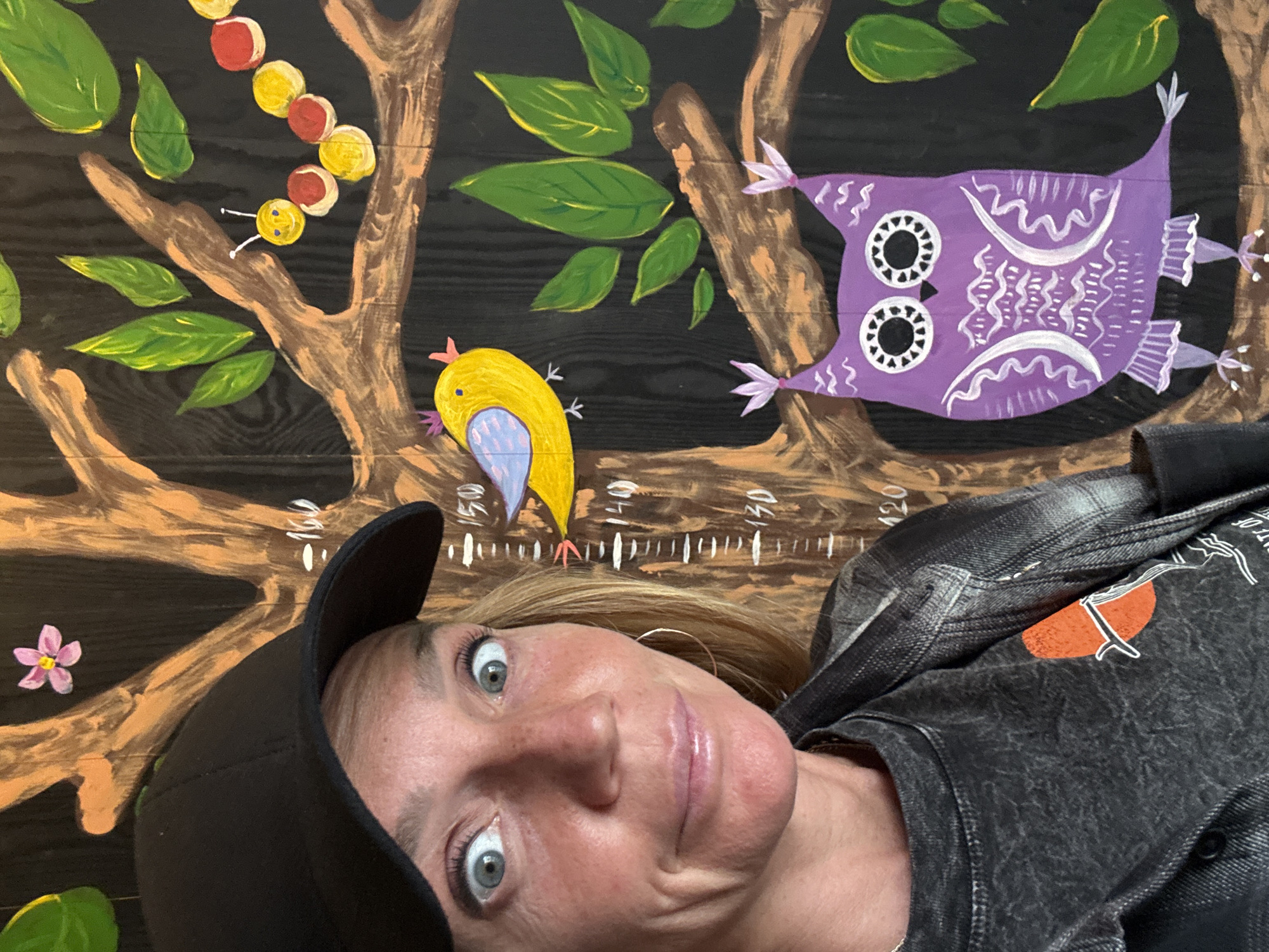 A woman with light-colored eyes and blond hair, wearing a black baseball cap, a dark grey t-shirt with a graphic print, and a grey and black plaid jacket, poses for a selfie in front of a mural painted on a dark wooden surface. The mural depicts a tree with brown branches and green leaves, a yellow bird with a blue wing, a colorful caterpillar, and a purple owl. A height chart is visible on the mural, with markings for 120, 130, 140, and 150 cm. The woman's expression is one of surprise, with wide-open eyes and slightly pursed lips. The lighting suggests an indoor setting, possibly a child's room or play area, though the specific location in "Unknown, Russia" cannot be determined from the visual information. The overall scene is informal and playful.