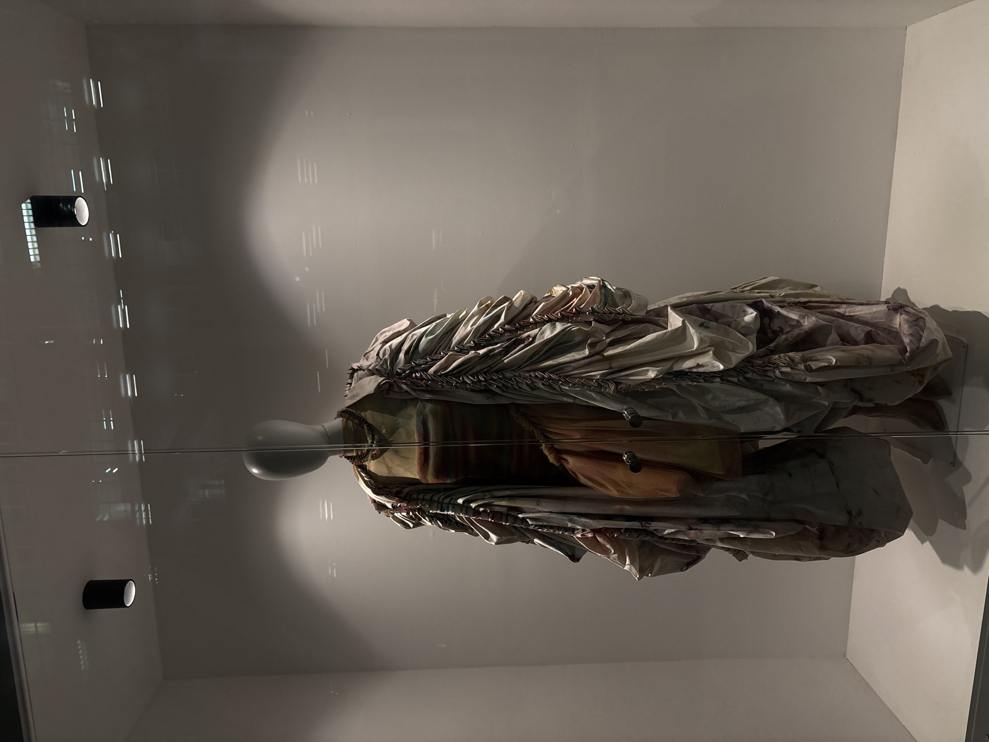 An elaborate, multi-layered garment is displayed on a mannequin within a clear glass case in what appears to be an exhibition or museum setting in Mexico City, Mexico. The image is oriented vertically, with the mannequin and dress lying horizontally across the frame.
The central subject is a white, featureless mannequin head and torso, presenting a voluminous, intricately designed dress or costume. The garment features numerous pleats, ruffles, and gathers, creating a textured and somewhat deconstructed appearance. The fabric tones are muted, predominantly shades of cream, beige, light brown, and pale grey, with hints of faded purplish and greenish hues, suggesting an aged or distressed aesthetic. Elements resembling cords or ties are visible along the upper edge of the garment.
The mannequin and dress are encased in a reflective glass display, which casts prominent reflections of the surrounding environment onto its surface. Two black, cylindrical lighting fixtures are visible mounted on the wall behind the display, one towards the top left and another towards the bottom left of the frame, and their reflections are clearly seen on the glass. Faint vertical lines, possibly from architectural features or windows in the room, are also reflected across the glass, interacting with the light sources. The background behind the display is a plain, light-colored wall, adding to the minimal and focused presentation of the exhibit. No visible text is present in the image.