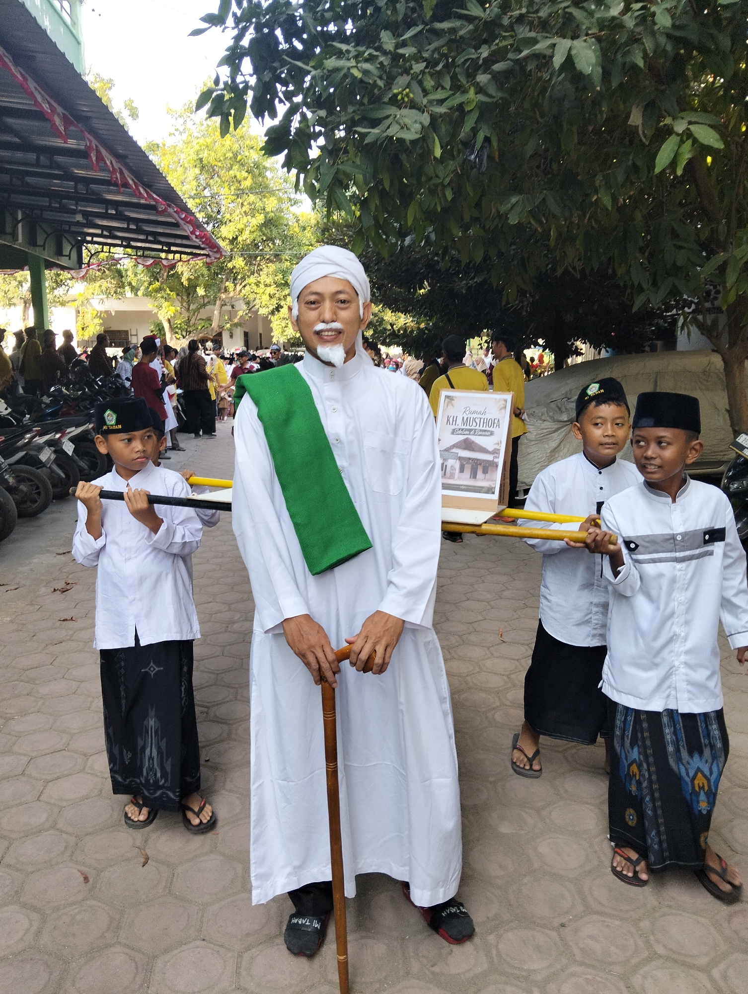 In the city of Kranji, Indonesia, a procession is underway under clear, sunny skies. The central figure, an adult male dressed in a white robe, white turban, and a green sash, walks with a wooden cane. He has a white mustache and goatee. Flanking him are two young boys, also in white robes and black caps. The boy on the left wears a black sarong with a pattern. The boy on the right wears a black sarong with a blue and yellow pattern. Both boys hold poles that support a wooden frame displaying a framed picture labeled "Rumah KH. Musthofa". Behind them, other adults and children are visible, some with motorcycles parked in the background. The ground is paved with hexagonal tiles. The scene is framed by trees with green foliage and a building with a red and white striped awning.
