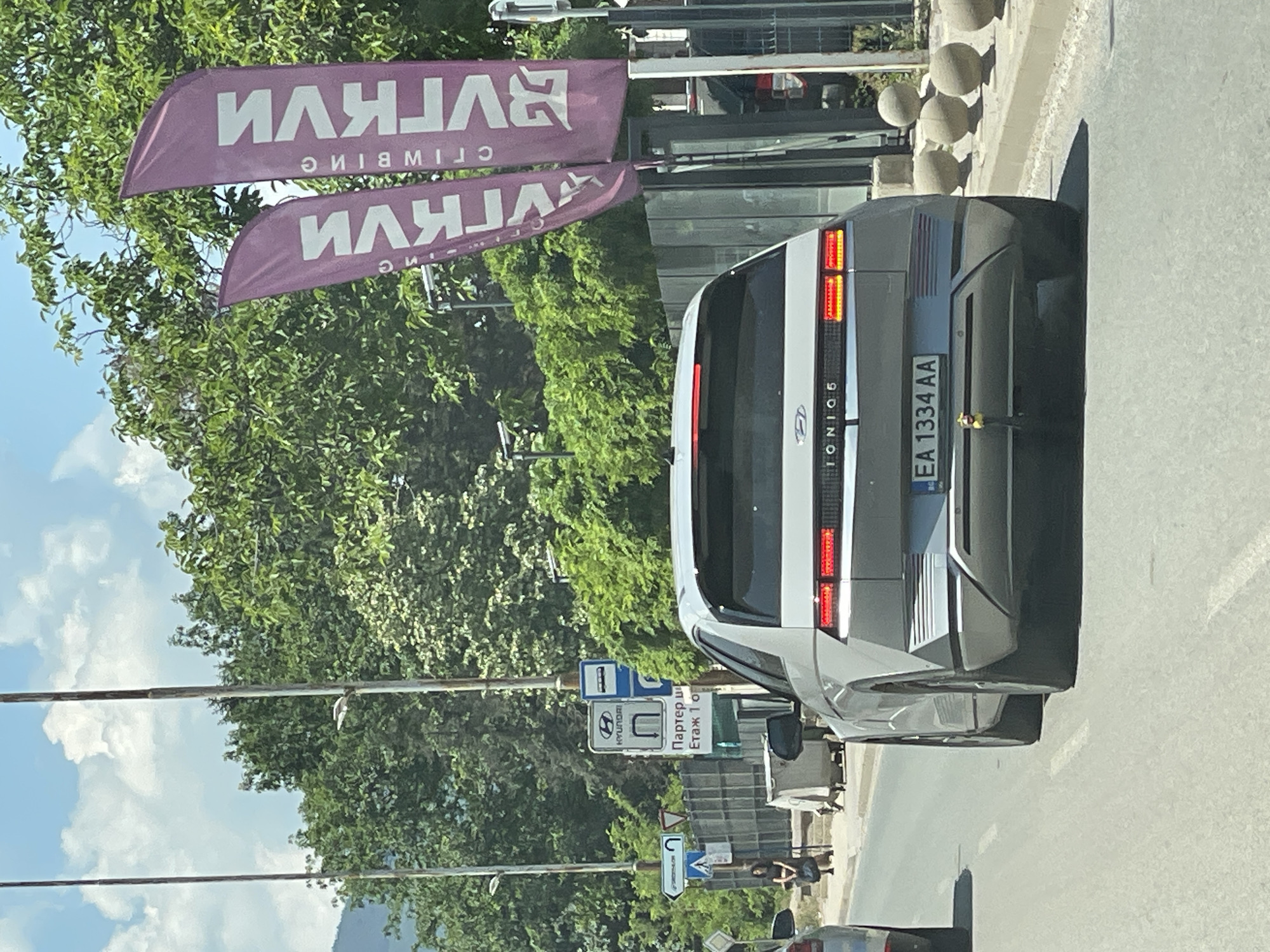 The image captures a rear view of a silver Hyundai Ioniq 5 electric vehicle driving on a paved road in an urban environment. The car has a distinctive modern design with horizontal LED taillights and the "IONIQ 5" badge visible. Its license plate, "EA 1334 AA," is clearly displayed. The car is in motion, indicated by the slight motion blur.
To the left of the car, a bus stop sign with a blue rectangle and a white bus icon is visible, accompanied by a sign indicating "Партер и Етаж 1 О," which translates to "Ground Floor and Floor 1." A Hyundai logo is also present on a vertical sign. Further left, a woman with dark hair and a dark top is standing near a trash receptacle.
In the background, lush green trees and foliage form a dense backdrop. Two tall, purple flags with white text, which appears to be Cyrillic, are prominently displayed on the right side of the image. The text on the flags, "ВИГКИ" and "ГКИ," likely refers to a brand or event. In the distance, a mountain range can be seen under a partly cloudy blue sky. The scene appears to be a sunny day.
Based on the presence of Cyrillic text on the signs and flags, the location is highly likely to be Bulgaria, specifically Sofia, given the commonality of this type of urban setting and signage. The road markings and general urban infrastructure are consistent with Bulgarian cities.
