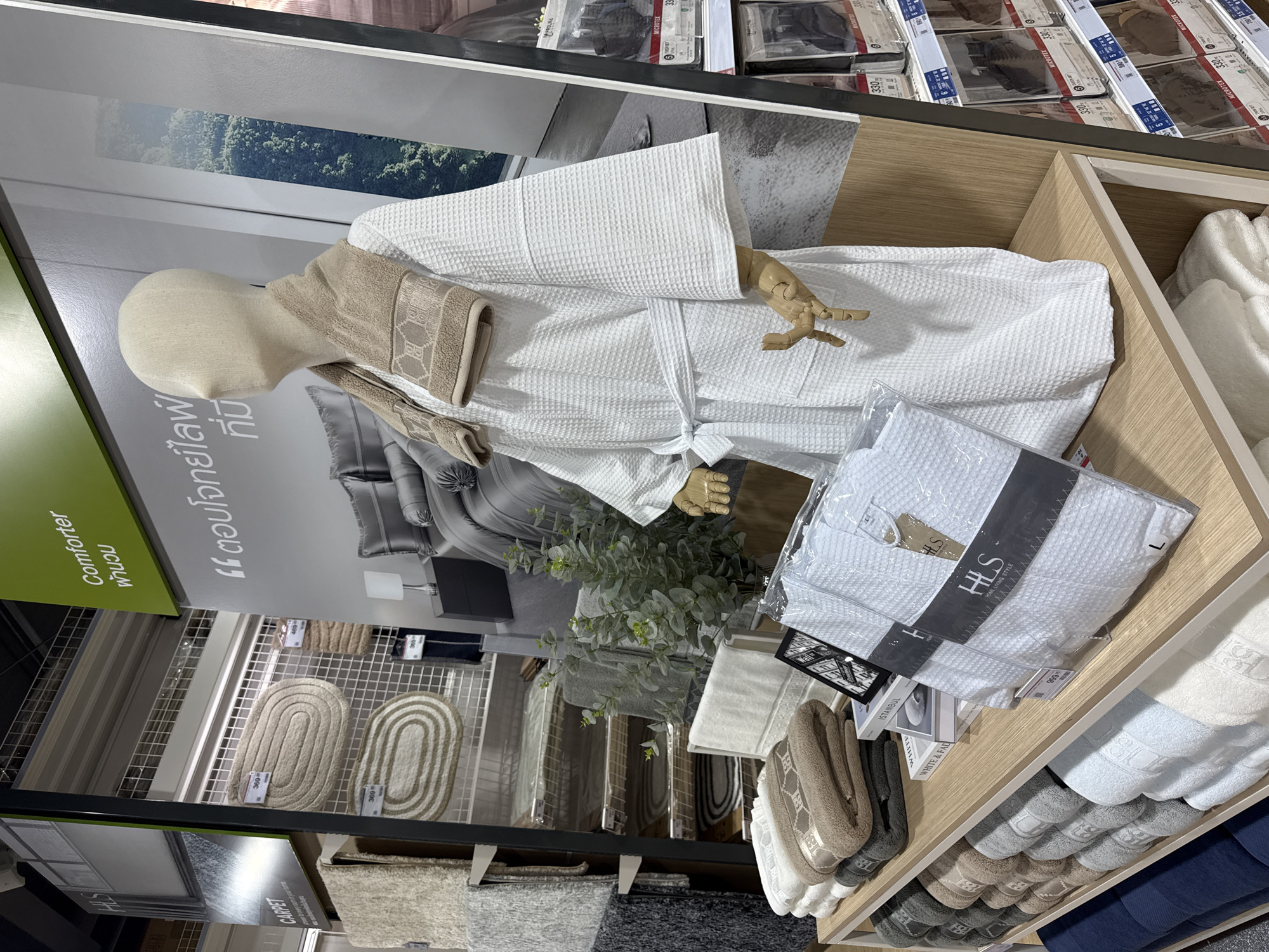 The image depicts a retail display of home textiles, likely in a department store or home goods store. The display features a mannequin dressed in a white waffle-knit robe, adorned with a beige towel draped around its neck. The mannequin stands in front of a large poster showcasing bedding and a lush green landscape.
On shelves to the left of the mannequin, various folded towels and bath mats are neatly stacked. These items are displayed in a range of colors, including beige, gray, white, and blue. Some of the towels have embroidered logos or patterns. Above these shelves, oval-shaped bath mats with textured surfaces are hung on a grid display. A sign reads "CARPET."
To the right of the mannequin, more shelves hold packaged bathrobes and towels. The packaging for the bathrobes includes the brand name "HLS HOME LIVING STYLE." Some of the shelves also feature price tags and smaller displays of packaged bedding sets.
The overall environment appears to be a well-lit retail space with a focus on creating an aspirational display for home furnishings. The presence of multiple product lines and organized presentation suggests a commercial setting. There are no discernible people or any specific details that would tie this image to the city of Baan Chaweng Noi, Thailand, beyond the requested location attribution which is not visually supported by the image content.