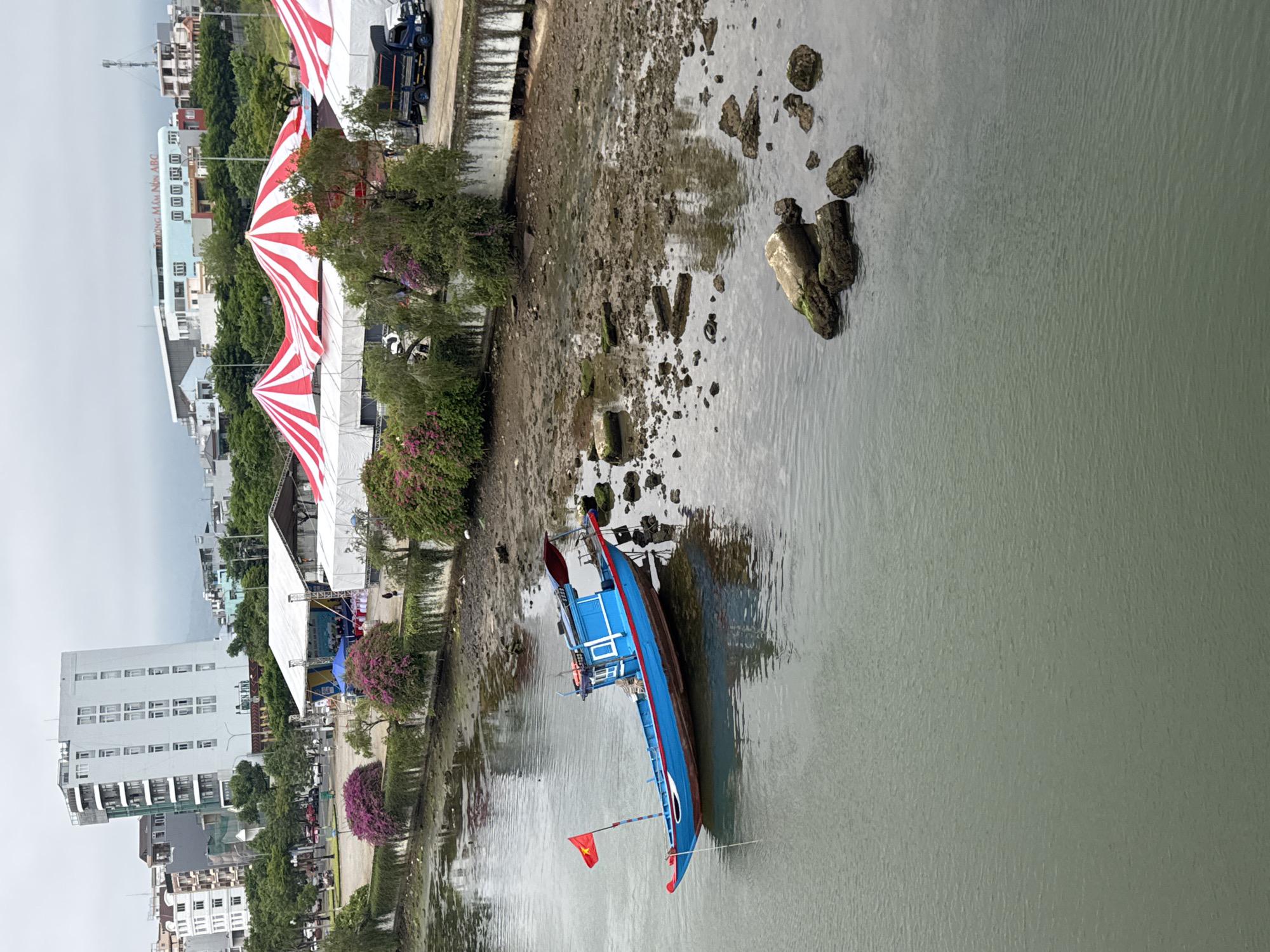 The scene is a waterway in Đà Nẵng, Vietnam, with a blue wooden fishing boat moored near the muddy bank. The boat, oriented diagonally in the frame, has a red Vietnamese flag attached. The riverbank shows signs of tidal fluctuation, with exposed mud and scattered rocks and debris. A row of trees and flowering plants lines the bank, beyond which is a paved area with tents decorated with red and white stripes, and a stage covered in white fabric. Beyond that are buildings of varying heights and architectural styles. The sky is overcast.
