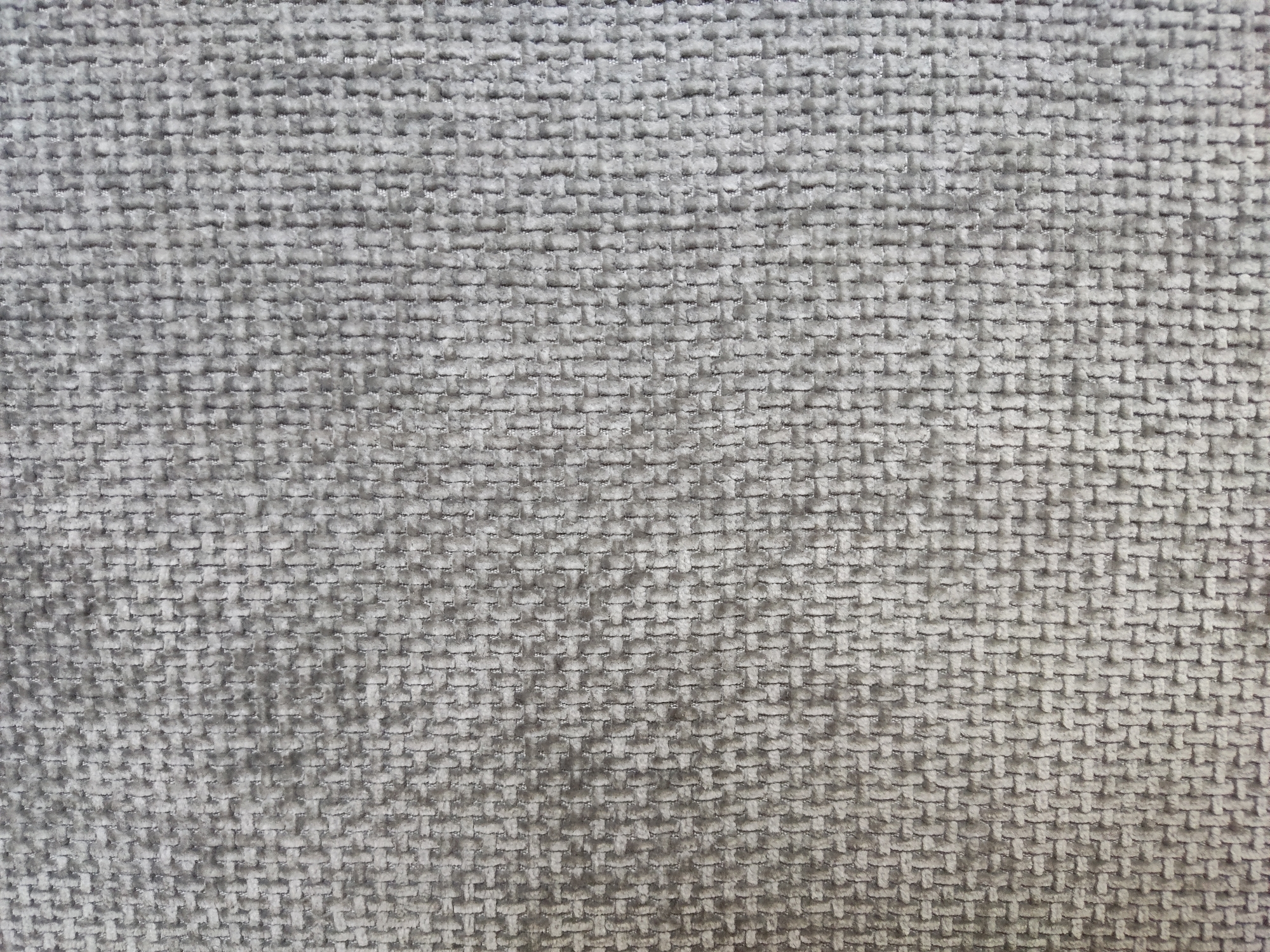 The image displays a close-up, full-frame view of a textured gray fabric. The fabric has a woven pattern, appearing to be a basketweave or similar interlocking weave structure. The surface of the fabric exhibits a slightly plush or velvety quality, with subtle variations in shading due to the texture and lighting, giving it a muted, soft appearance. The color is a consistent medium gray. No people, specific objects, or environmental context beyond the fabric itself are visible. This image is a detailed texture sample. There is no discernible information related to the city of Бараўлянскі сельскі Савет, Belarus within the image.