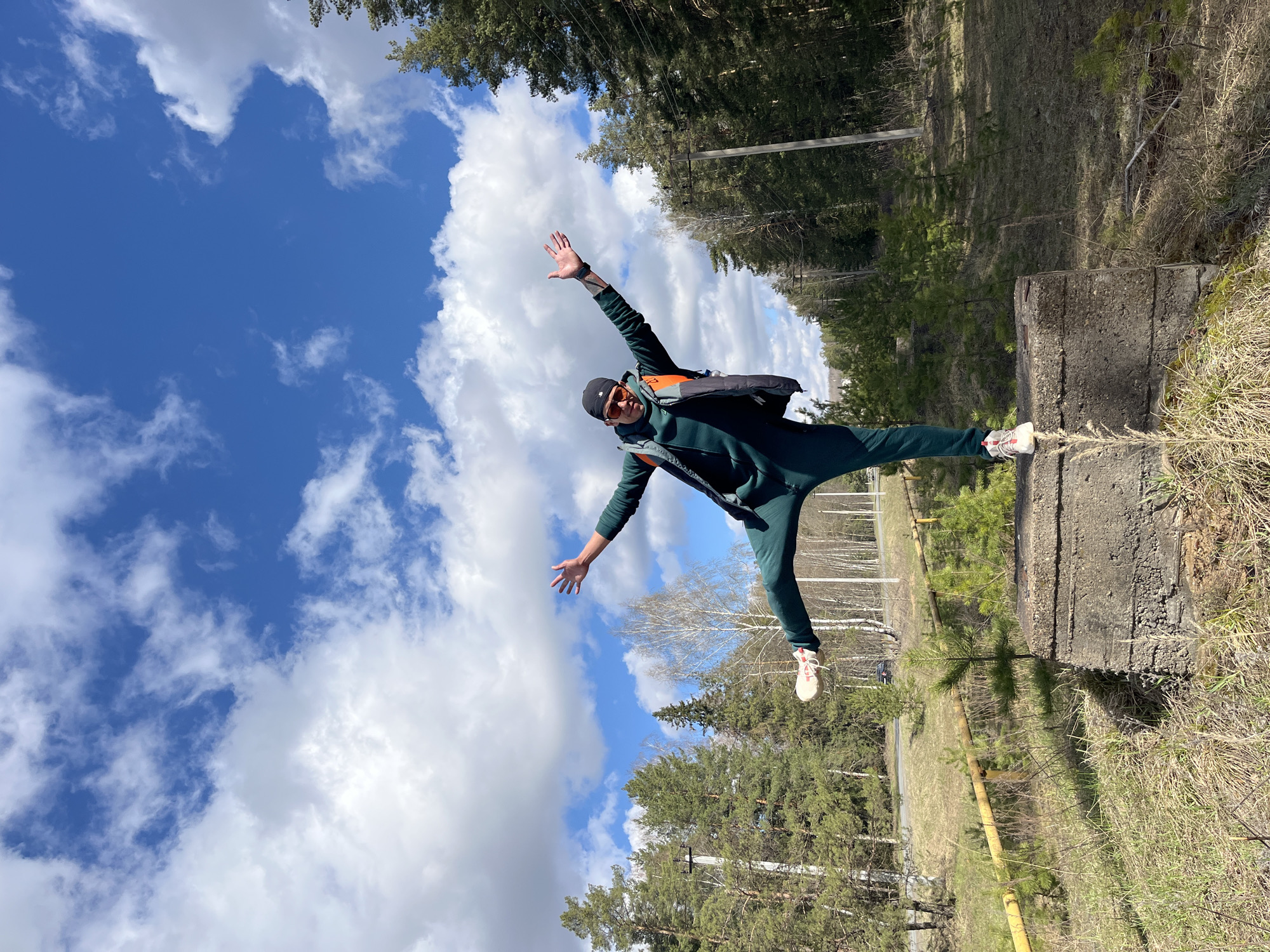 This outdoor image, taken in Noviy, Russia, features a person captured in a dynamic pose against a clear blue sky with scattered white clouds. The subject, a male individual, is prominently featured in the center-right of the frame, balancing on one leg atop a weathered, dark grey concrete block. He is dressed in a dark green tracksuit, a black beanie, and orange-tinted sunglasses, and also wears a backpack or vest with an orange section. His arms are outstretched, conveying a sense of balance, joy, or freedom, with a tattoo visible on his left forearm. He is wearing white and red athletic shoes.
The setting is a natural or semi-natural landscape, likely a forest edge or rural area. Tall coniferous trees form a dense backdrop, particularly on the left side, interspersed with some bare deciduous trees. The ground is covered with dry grass and sparse vegetation. In the mid-ground, a yellow pipe or barrier runs horizontally across the landscape, and a utility pole with overhead lines is visible. A dark vehicle is partially discernible on a distant road.
The bright sunlight and clear sky with fluffy cumulus clouds indicate fair weather, likely midday or early afternoon. The overall impression is one of a cheerful and active individual enjoying the outdoors. No visible text is present in the image.