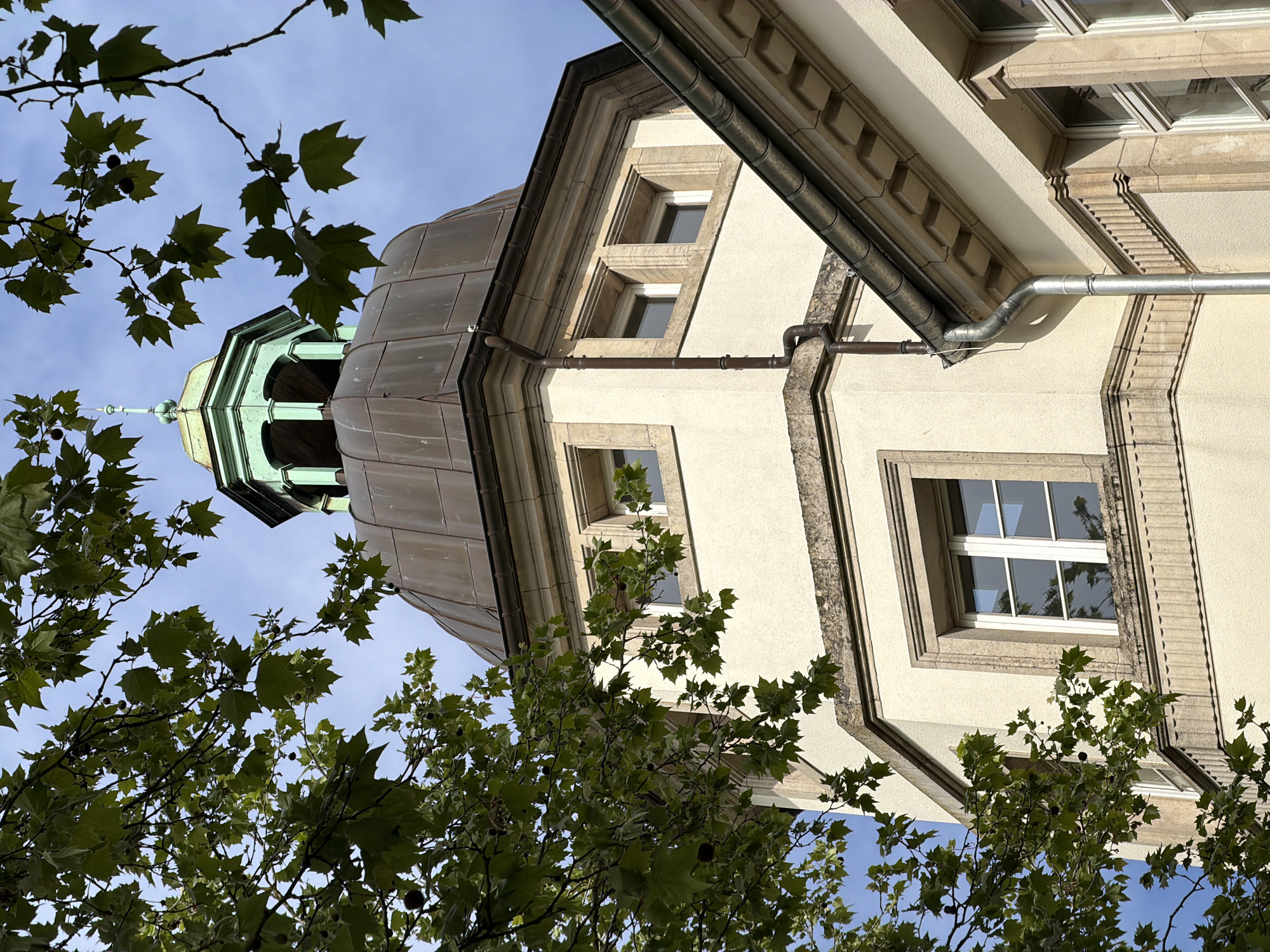 The image depicts an upward angle view of a building in Esch-sur-Alzette, Luxembourg. The structure features a tower with a copper-colored dome that transitions to a green cupola at the top. A spire-like element crowns the cupola. Parts of the building facade are visible, exhibiting a cream-colored stucco with light-colored stone framing windows. A metal gutter system runs along the edge of the building. The sky is visible in the background, with a blue hue interspersed with some cloudiness. Tree branches and leaves partially obscure the view, adding a natural foreground element to the architectural scene.