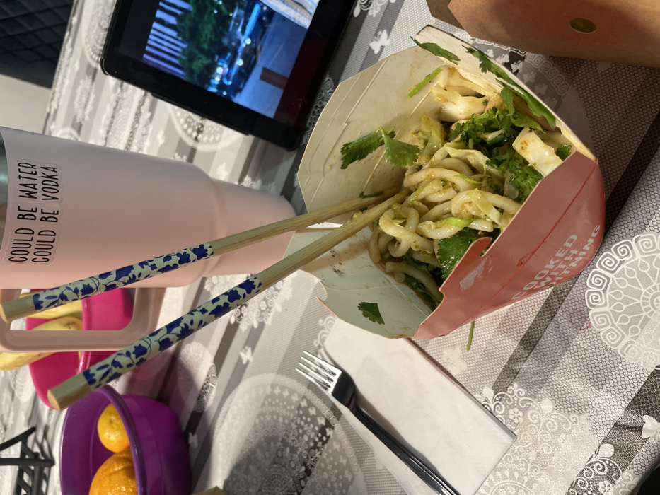 A tabletop scene reveals a box of noodles with cilantro, accompanied by chopsticks. A pink tumbler with the quote "could be water, could be vodka" sits nearby. A tablet displays a video, and a fork rests on a napkin. A purple bowl containing oranges is visible in the corner. The table is covered with a patterned tablecloth.