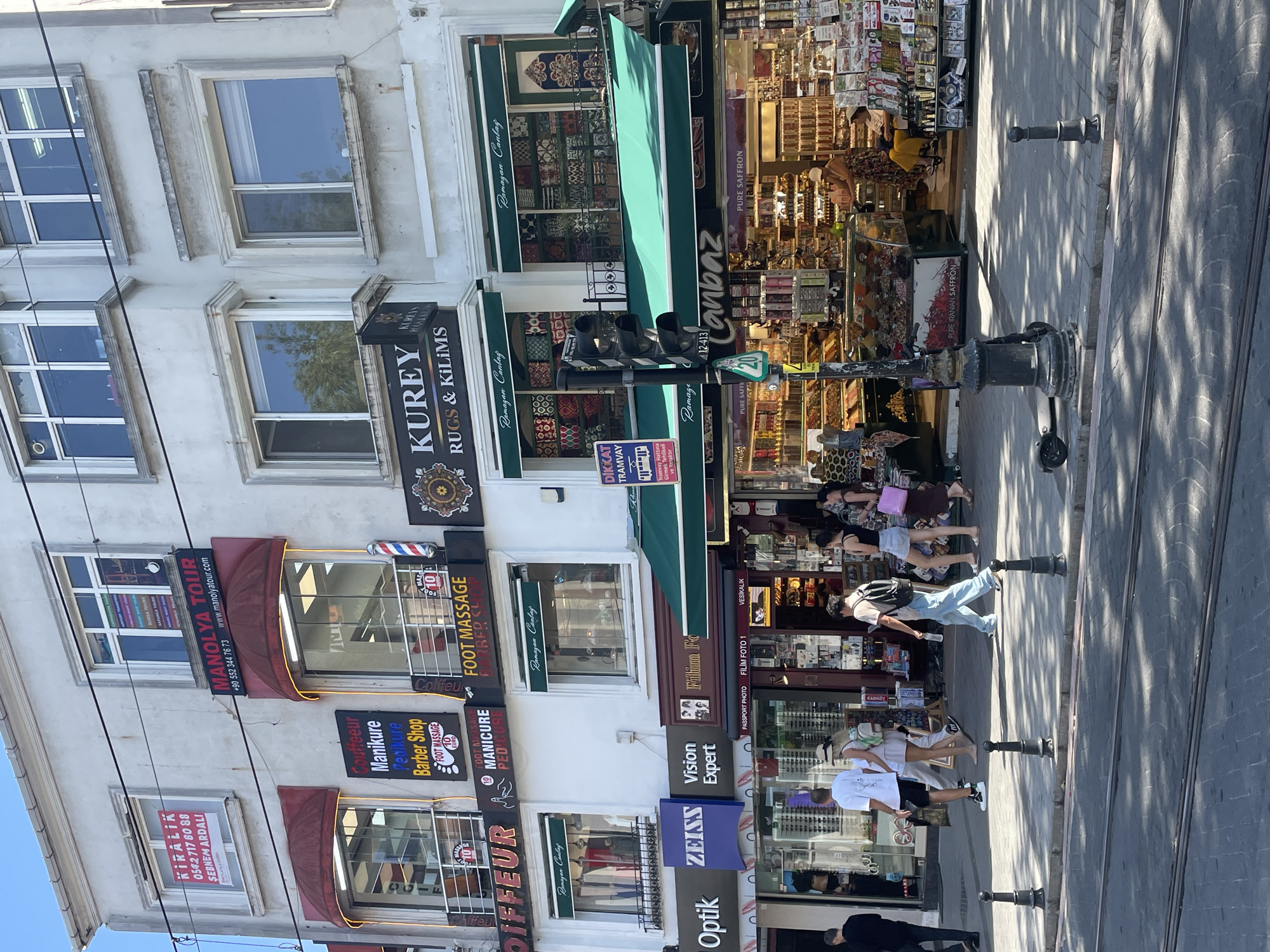 The image captures a street scene in Istanbul, Turkey, likely on a sunny day given the strong shadows. The foreground features tram tracks on a paved street, with bollards lining the sidewalk. In the mid-ground, pedestrians are walking along the sidewalk in front of a row of shops. One person is wearing a white t-shirt and shorts, another in a light-colored dress, and a person with a backpack is walking towards the right. The shops themselves are varied, with awnings and signs indicating businesses such as "Optik," "ZEISS Vision Expert," and "Filhima Fo." Further up the building, signs for "MANOLYATOUR," "Couffeur Manicure Pedicure Barber Shop," and "FOOT MASSAGE" are visible. Another shop displays "KUREY RUGS & KILIMS." The shops sell a variety of goods, including what appear to be spices, saffron, and carpets, as evidenced by displays in the windows and on shelves. The architecture is multi-story, with multiple windows and awnings. A traffic light stands on the sidewalk. The overall impression is a bustling commercial street in a Turkish city.