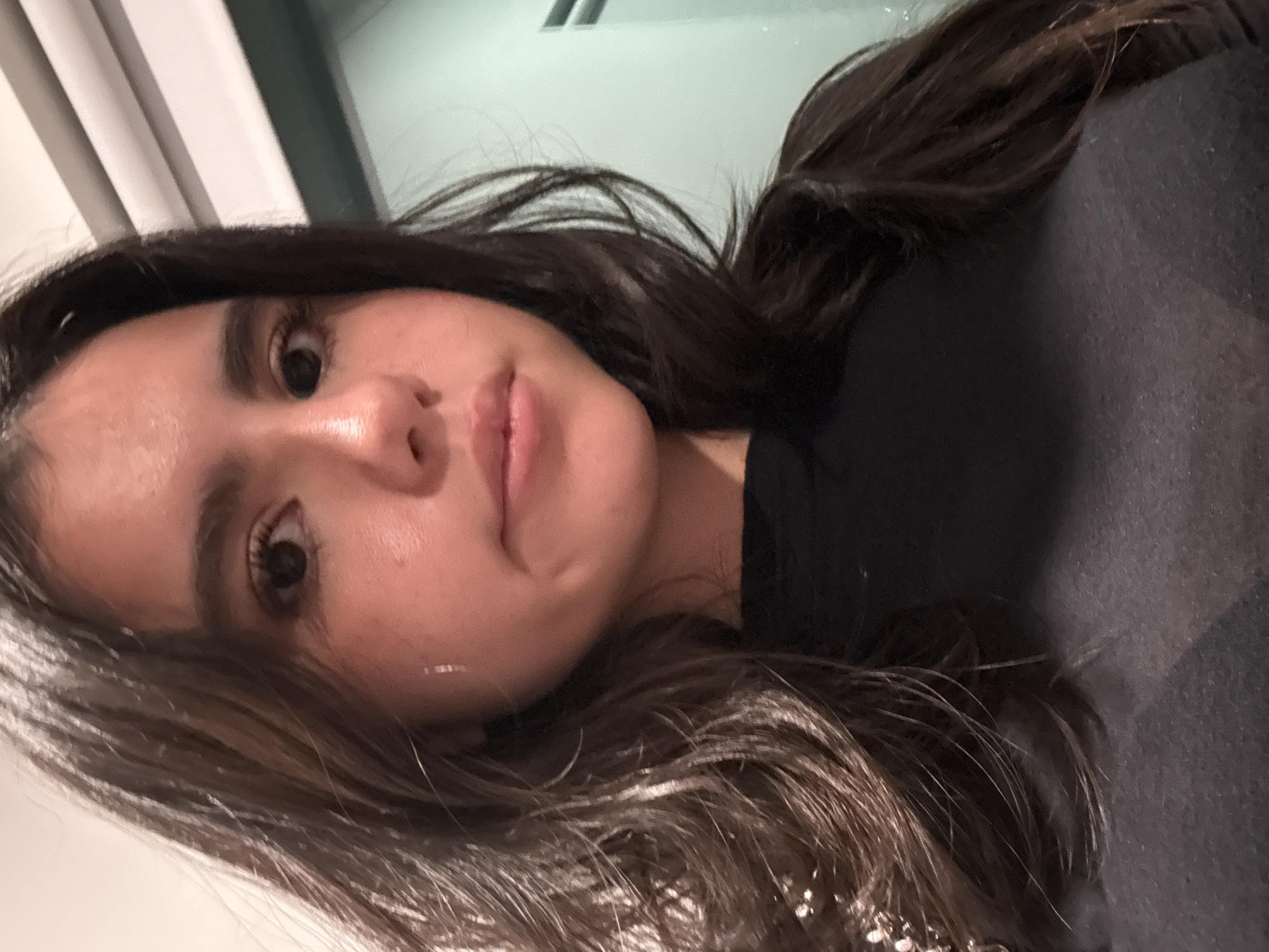 A close-up portrait, rotated 90 degrees counter-clockwise, features a young woman in an indoor setting in Austin, United States of America. Her head is tilted to the right, and she looks directly at the camera with a neutral to subtly pensive expression. She has long, dark brown hair that frames her face and falls over her left shoulder. Her eyes are dark and appear slightly wide, with visible dark eyelashes and eyebrows. Her lips are full and slightly parted, showing a natural pinkish tone. A distinct bright highlight or reflection, resembling a tear, is visible on her left cheek. She is wearing a dark-colored top, likely black or dark grey, visible on her upper body. The background is out of focus, revealing light-colored, possibly paneled, surfaces, suggesting an indoor environment with soft or artificial lighting. No text is visible in the image.