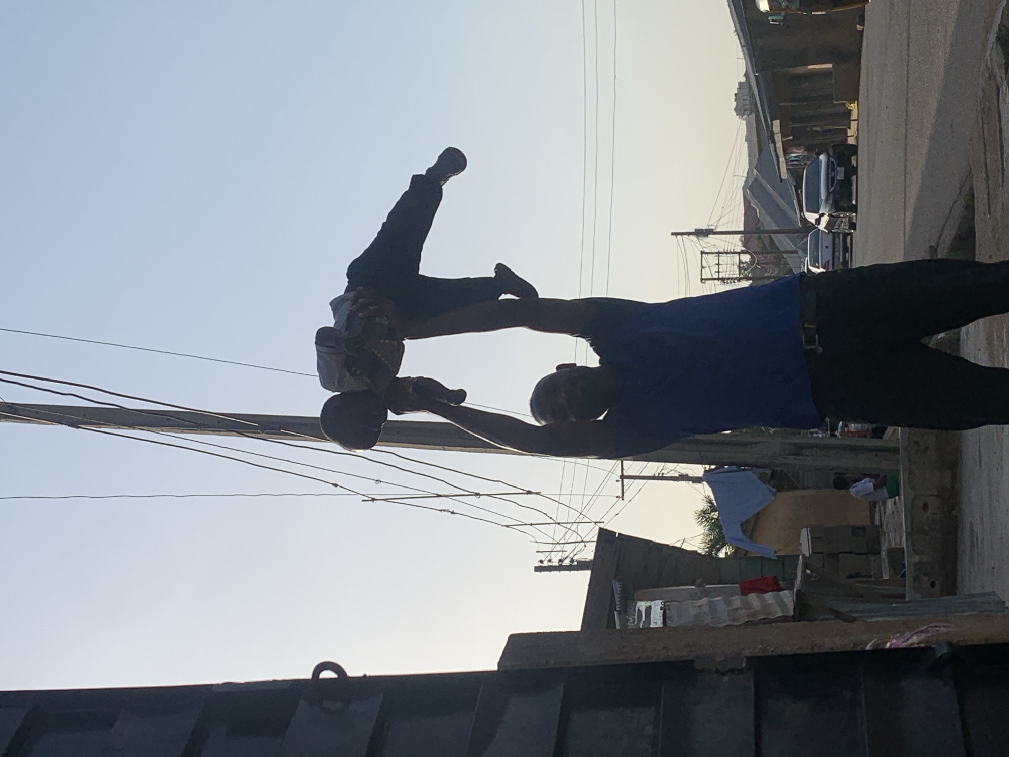 The image shows a man lifting a child in the air in what appears to be a residential area. The man is wearing a blue t-shirt and dark pants, and the child is wearing a light-colored shirt and dark pants. They are positioned in the foreground, with the man's arms extended upwards to hold the child.
In the background, there are buildings, some of which appear to be made of corrugated metal and concrete. There are also power lines running overhead and a few cars parked along a street. The sky is clear and light blue, suggesting it is daytime. The overall environment looks like a busy street in a developing urban area.
While the image itself does not explicitly state the location, based on the style of buildings, vehicles, and clothing, it is plausible that this scene is from the city of Jos, Nigeria. Jos is known for its diverse population and vibrant street life, which would be consistent with the visual elements present in the photograph.