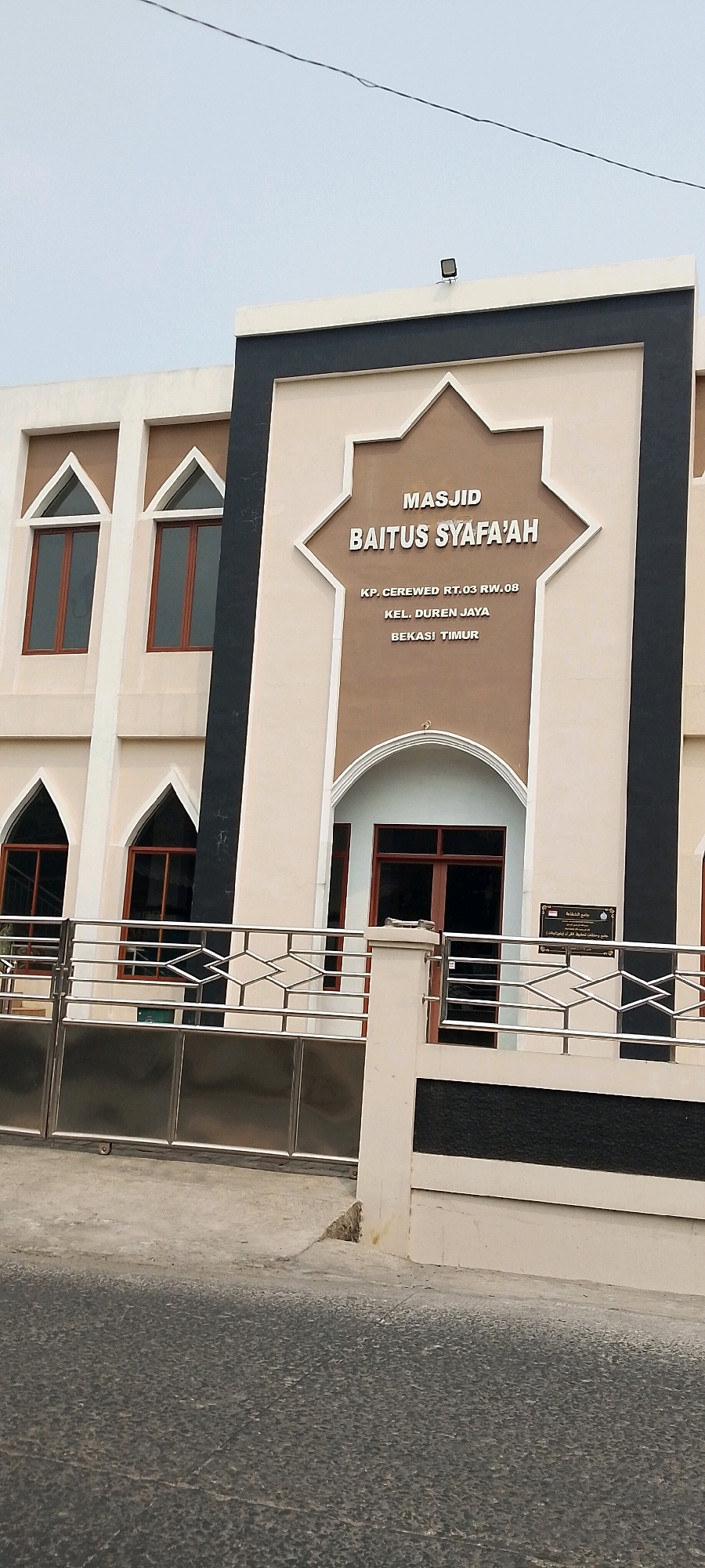 The image shows the facade of a mosque named "MASJID BAITUS SYAFA'AH" in Bekasi, Indonesia. The mosque has a modern architectural style with beige and dark grey accents. The name of the mosque is prominently displayed in raised silver lettering. Below the mosque's name, the address is inscribed: "KP. CEREWED RT.03 RW.08", "KEL. DUREN JAYA", and "BEKASI TIMUR". The building features several arched windows with dark frames, and a main entrance with a similar arched design. In front of the building, there is a stainless steel gate and fence. The ground in front of the mosque is paved with grey asphalt. The sky is overcast.