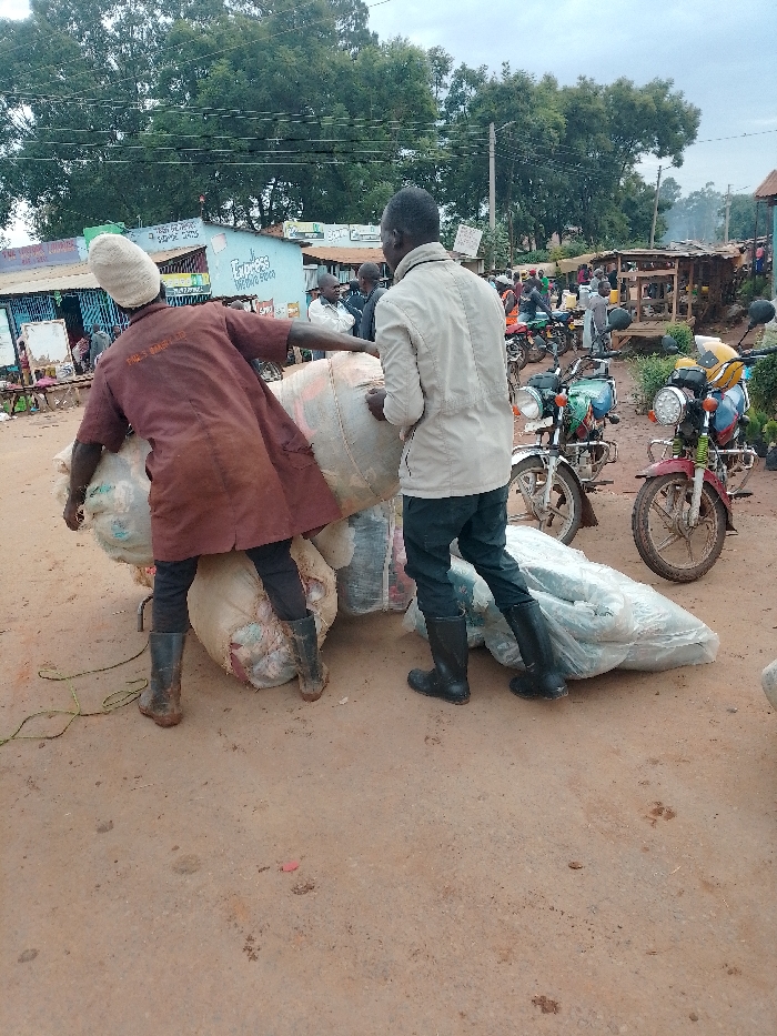 The image shows two men loading large, wrapped bundles onto a motorcycle. The men are standing on a dirt road in front of a small store, and the road is lined with other motorcycles and motorbikes. The man in the foreground, wearing a brown jacket, is lifting a large bundle onto the motorcycle. The man behind him, wearing a beige jacket, is helping to secure the bundle. The background is a dense urban area with multiple structures and vehicles, some in motion. The scene appears to be a typical trading activity in the busy area of Kivaywa, Kenya, located at coordinates 0.562 latitude and 34.805 longitude. The scene suggests a bustling marketplace with commerce and trade occurring.