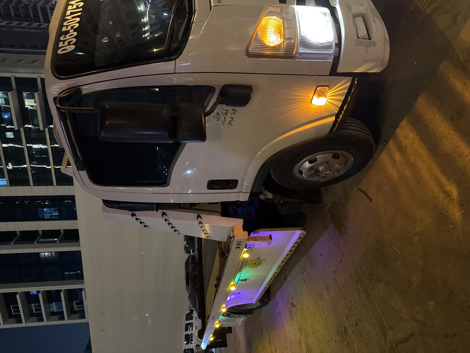 A close-up of a white tow truck angled diagonally. The front section showcases the truck's cabin, headlight, and front wheel. Reflections of a modern building are visible in the cabin window, indicating an urban setting at night. The towing platform, extending from the truck, features embedded lights. Part of the truck has Arabic writing. The truck is likely stationary.