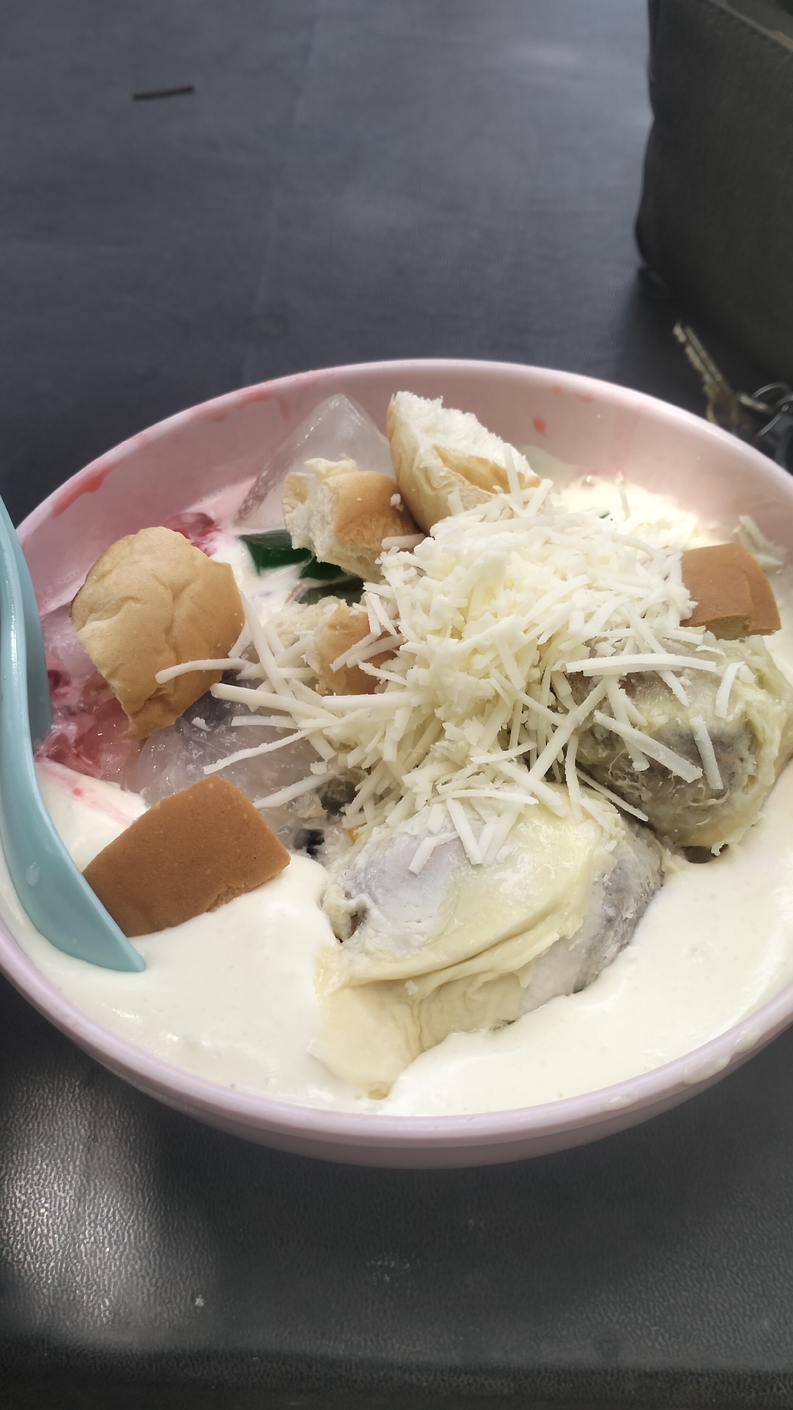 In Lhokseumawe, Indonesia, a light pink bowl contains a dessert. The bowl is filled with a creamy white liquid, ice shards, chunks of bread, green jelly, and pieces of durian fruit. The dessert is topped with shredded white cheese. A light blue spoon is partially submerged in the left side of the bowl. The bowl rests on a dark, textured surface, possibly a table. In the background, a dark bag and keys are visible.