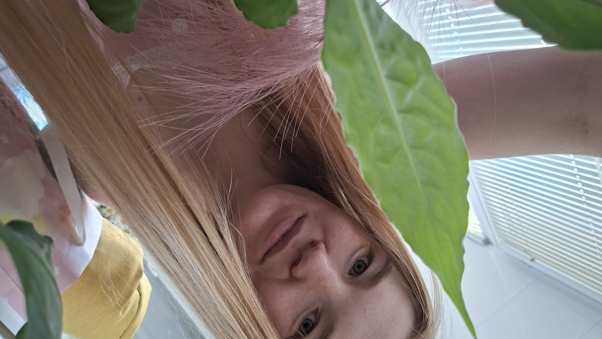 A young woman with blonde hair and freckles is taking a selfie, holding a green leaf in the foreground. She is wearing a pink, feathery top. Behind her, there are horizontal blinds on a window, and parts of yellow and pink fabric are visible, possibly clothing or decorative items. The image appears to be taken indoors, with natural light coming from the window. There is no indication of the location being in Chaikovsky, Russia.