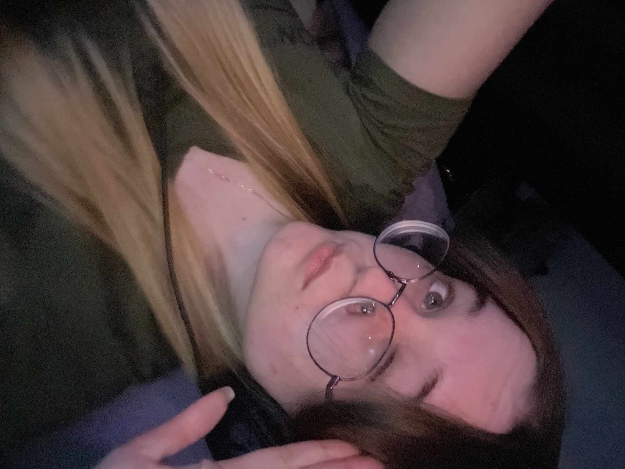 This image presents a close-up, angled portrait of a young female subject located in the city of Pushchino, Russia. The subject has long hair, dark brown at the roots and fading to a lighter blonde or ombré towards the ends, which falls over her shoulders and chest. She is wearing round, wire-rimmed eyeglasses and a dark olive green short-sleeved t-shirt. A thin silver-colored chain necklace is visible around her neck. A black headset is worn on her head, partially obscured by her hair and her raised left hand. Her fingers are positioned near her forehead, seemingly adjusting the headset or her hair. Her face is illuminated by a predominant reddish-pink light, highlighting her fair complexion. Her eyes are light-colored, and she maintains direct eye contact with the camera. Small dermal marks are present on her left cheek. The background is dark and out of focus, indicating an indoor environment with low ambient lighting.