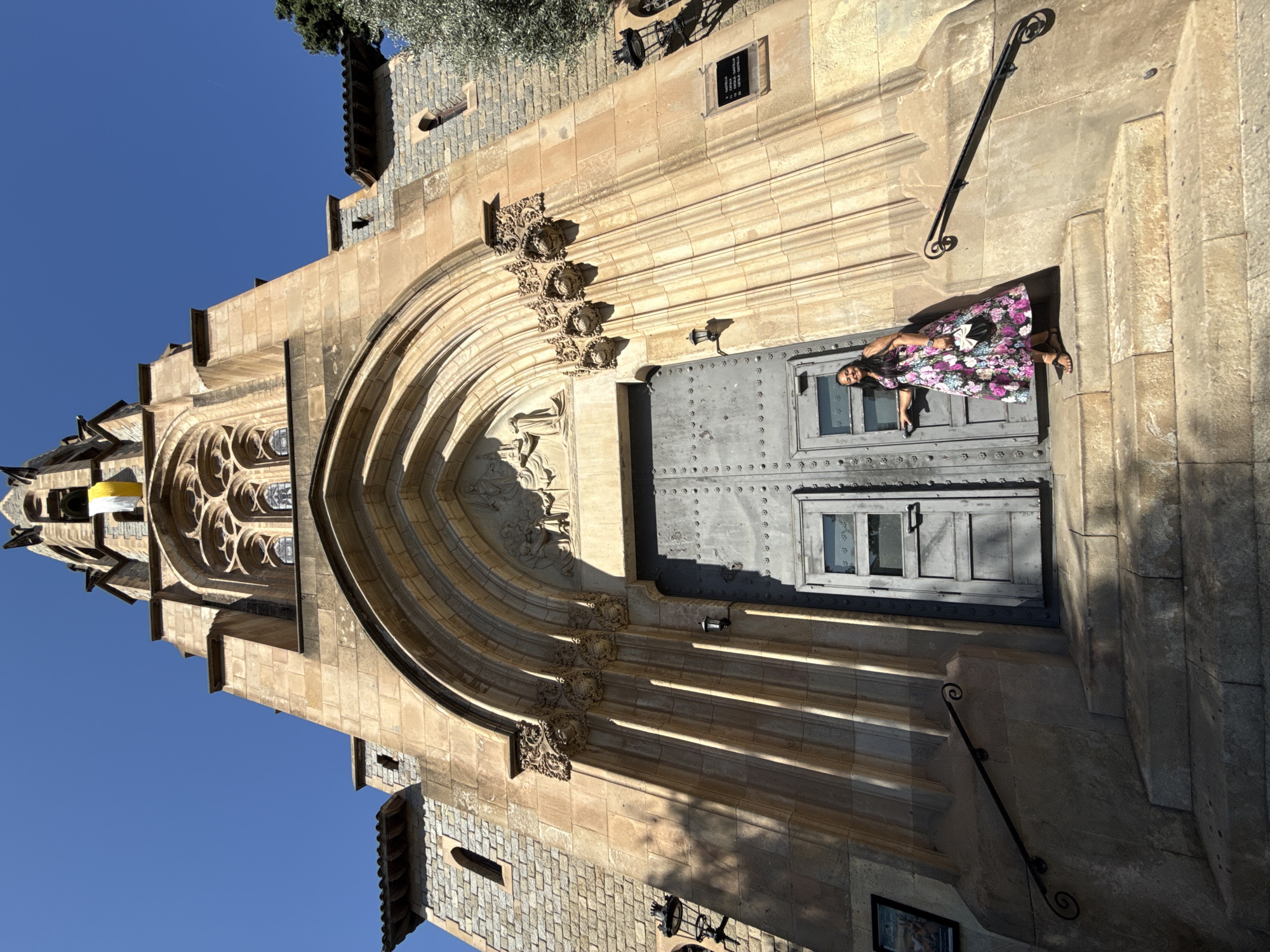 The image shows a low-angle, full-shot view of a woman standing on the stone steps in front of a large, ornate church entrance. The church is constructed of light-colored stone and features a tall, Gothic-style tower, arched doorways, and decorative carvings. A yellow and white flag hangs from the tower. The woman, wearing a floral print dress and holding a small black purse, is standing to the right of the double doors, with her right hand on one of the door handles. The steps are wide and made of rough-hewn stone, with a black metal railing on the right side. The sky above is clear and blue. This is the Church of Sant Vicenç in Santa Coloma de Gramenet, Spain.