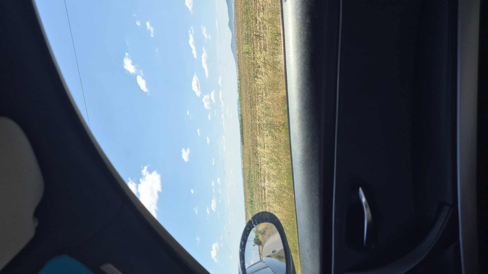 The image is a view from the interior of a vehicle, looking out the passenger side window. The foreground is dominated by the dark interior of the car door, including a silver door handle. Through the window, a vast, flat landscape stretches to the horizon under a bright blue sky dotted with fluffy white clouds. The landscape appears to be agricultural fields, with patches of dry grass and some cultivated rows. A curved road is visible in the reflection of the side-view mirror. The overall impression is of a rural setting on a sunny day, viewed from a moving car. The location is Unknown, Unknown.