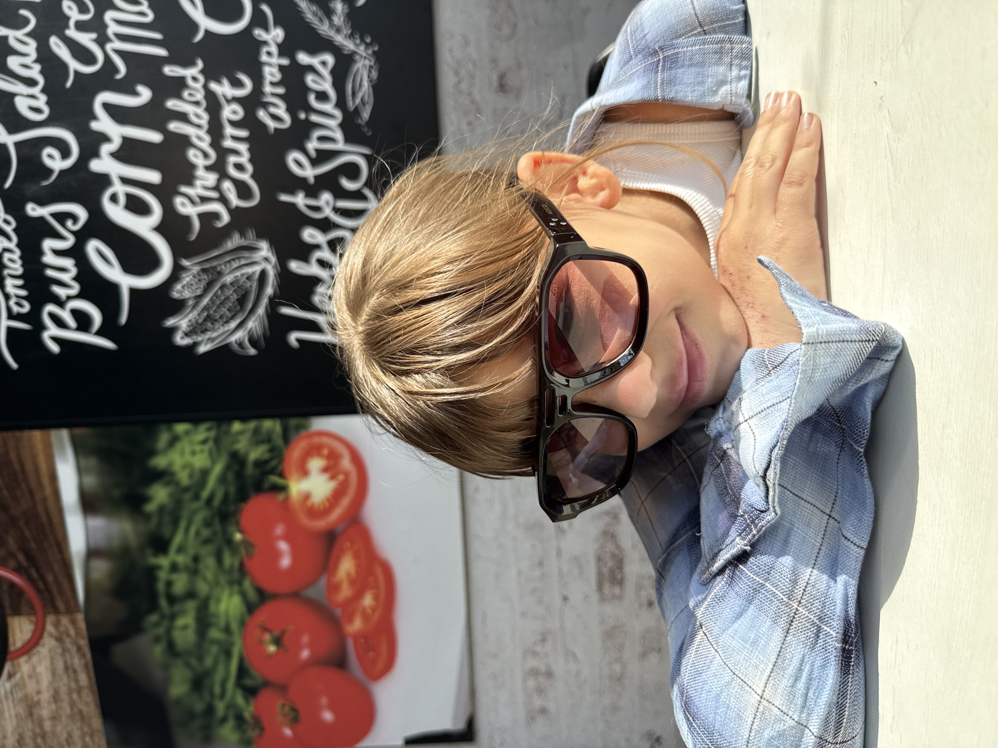 A child wearing sunglasses and a plaid shirt is lying on a white surface next to a sign. The sign advertises tomato salad, buns, corn, shredded carrot wraps, and herbs & spices. The sign also features illustrations of tomatoes and a grain stalk. The location is not discernible as being Шебунино, Russia.
