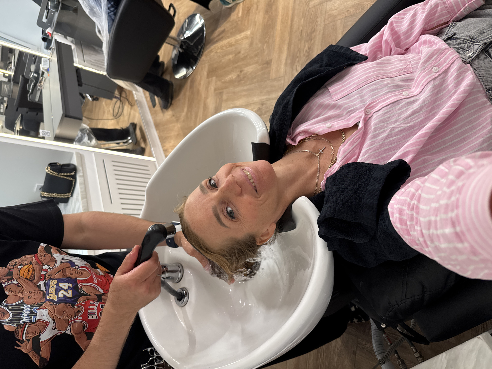 A fair-skinned woman with blond hair is reclining in a black salon chair with a black towel draped around her neck. She is wearing a pink and white striped shirt and a gold chain necklace with a cross pendant. Her head rests in a white salon sink while an unseen person washes her hair with a black handheld showerhead. A dark-skinned person's arm and hand are visible, holding the showerhead, and the person is wearing a black T-shirt with basketball player graphics. The salon appears to have light brown wood-look flooring. In the background, there are salon chairs, styling stations, and a black purse with a chain strap is visible. The city of Saint Petersburg, Russia cannot be confirmed based on the image.