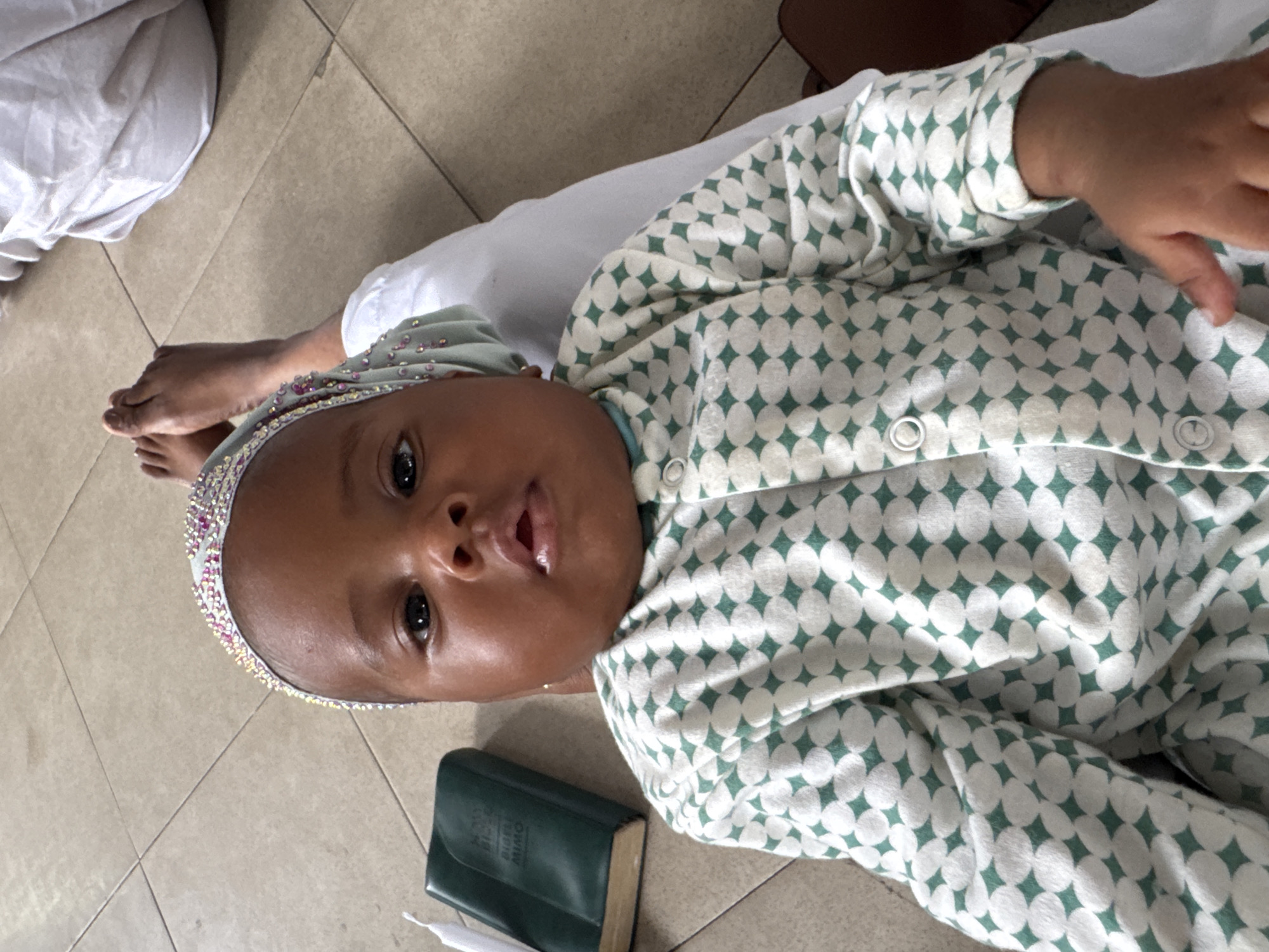 A baby wearing a white and green patterned outfit and a bejeweled head wrap is lying on a tiled floor. Next to the baby is a small, dark green book and a white object. The location is presumed to be in Badagry, Nigeria.