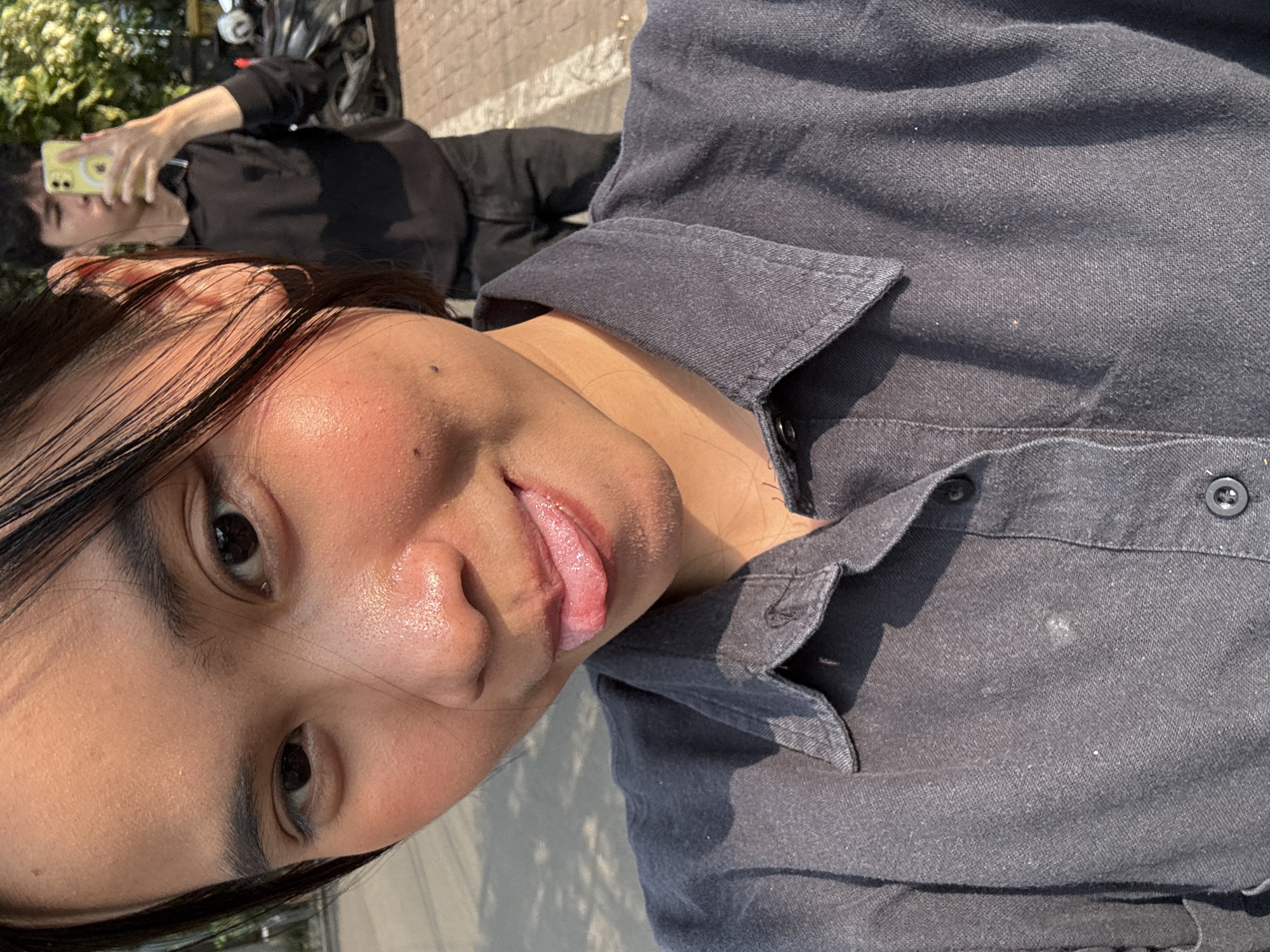 The media file shows a close-up, low-angle selfie of a person wearing a dark grey, collared shirt. The person's face is partially visible, with their tongue sticking out. Their skin appears shiny, suggesting it might be warm or humid. To the left of the main subject, another person is visible in the background, holding a yellow smartphone and seemingly taking a photo. This second person is wearing a black hoodie and dark pants.
In the background, a paved area with a white line is visible, along with some greenery and the side of a motorcycle. The sunlight suggests it is daytime, likely with clear weather. The shadows cast on the ground indicate a bright sun.
The setting appears to be outdoors in a place with some urban elements, given the paved ground and motorcycle. The overall impression is casual and relaxed. The activity appears to be taking a selfie, with another person also engaged in taking a photo. The time of day is likely midday due to the strong sunlight and shadows. The emotions conveyed are playful and lighthearted due to the tongue-out pose. No visible text is present in the image. The location context is South Jakarta, Indonesia.