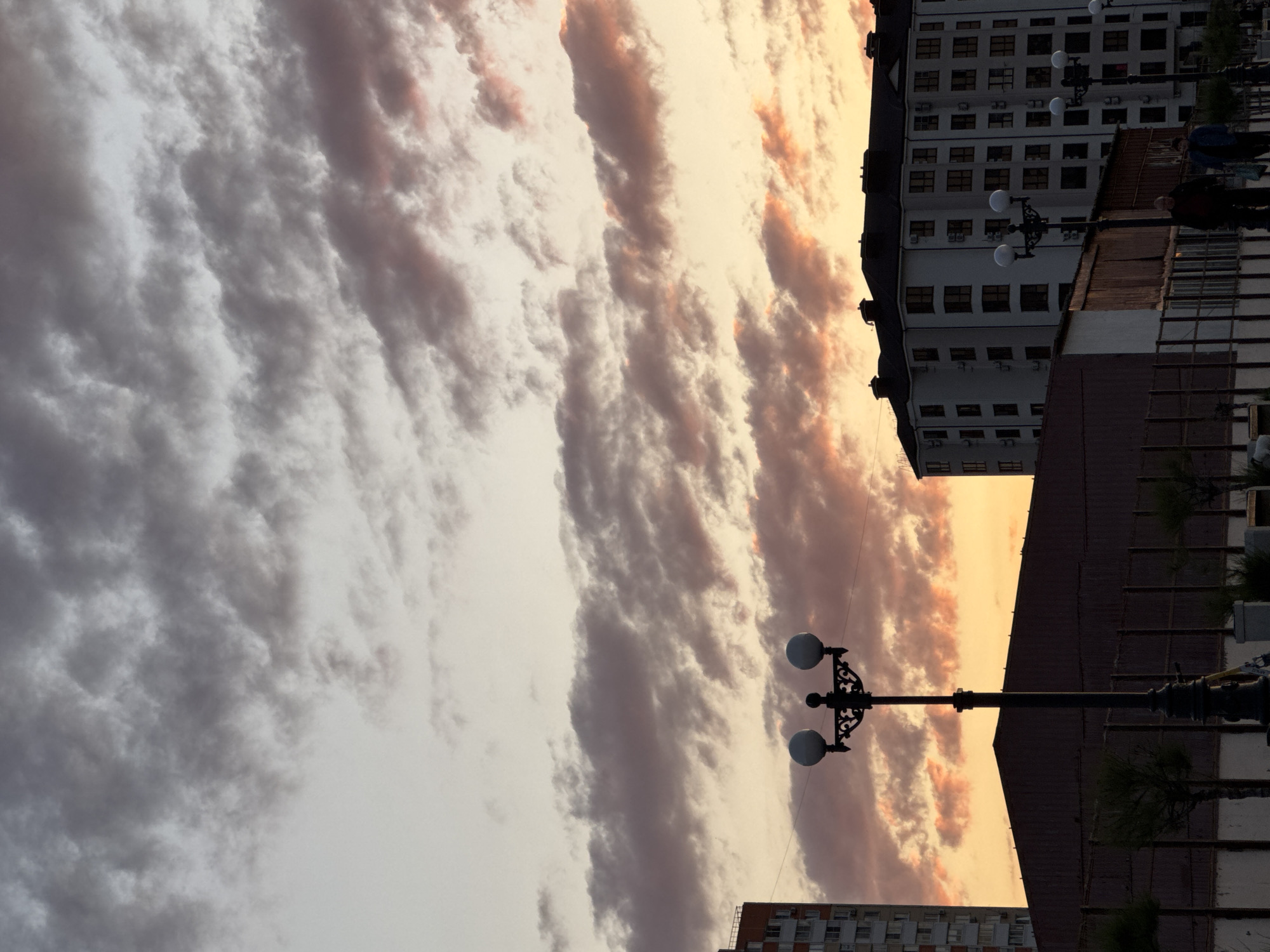 This image captures an urban twilight scene in Novorossiysk, Russia, dominated by a dramatic sky and city silhouettes. The sky is filled with elongated, textured clouds, possibly altocumulus or stratocumulus undulatus, exhibiting a striking gradient from light grey and white on the left to warm orange, pink, and yellow hues on the right, strongly suggesting either sunrise or sunset.
In the foreground and midground, two ornate black lampposts, each topped with two spherical white lights, stand tall, creating dark silhouettes against the bright sky. On the right side of the frame, several buildings are visible. One prominent multi-story building is light-colored with many windows and visible air conditioning units. Below this, a darker, possibly industrial-style building or wall stretches horizontally across the lower mid-section of the image, with small trees or bushes barely visible.
Two people are discernible in the midground, near one of the lampposts and buildings; one wears a blue jacket, and the other a darker outfit, appearing to be walking or standing. The activity is limited to a candid moment of people in an urban setting. The notable details include the vibrant colors of the sky contrasting with the dark urban elements, and the distinct cloud formations. No visible text is present. The overall mood is peaceful and visually impactful, showcasing the atmospheric conditions over the city.