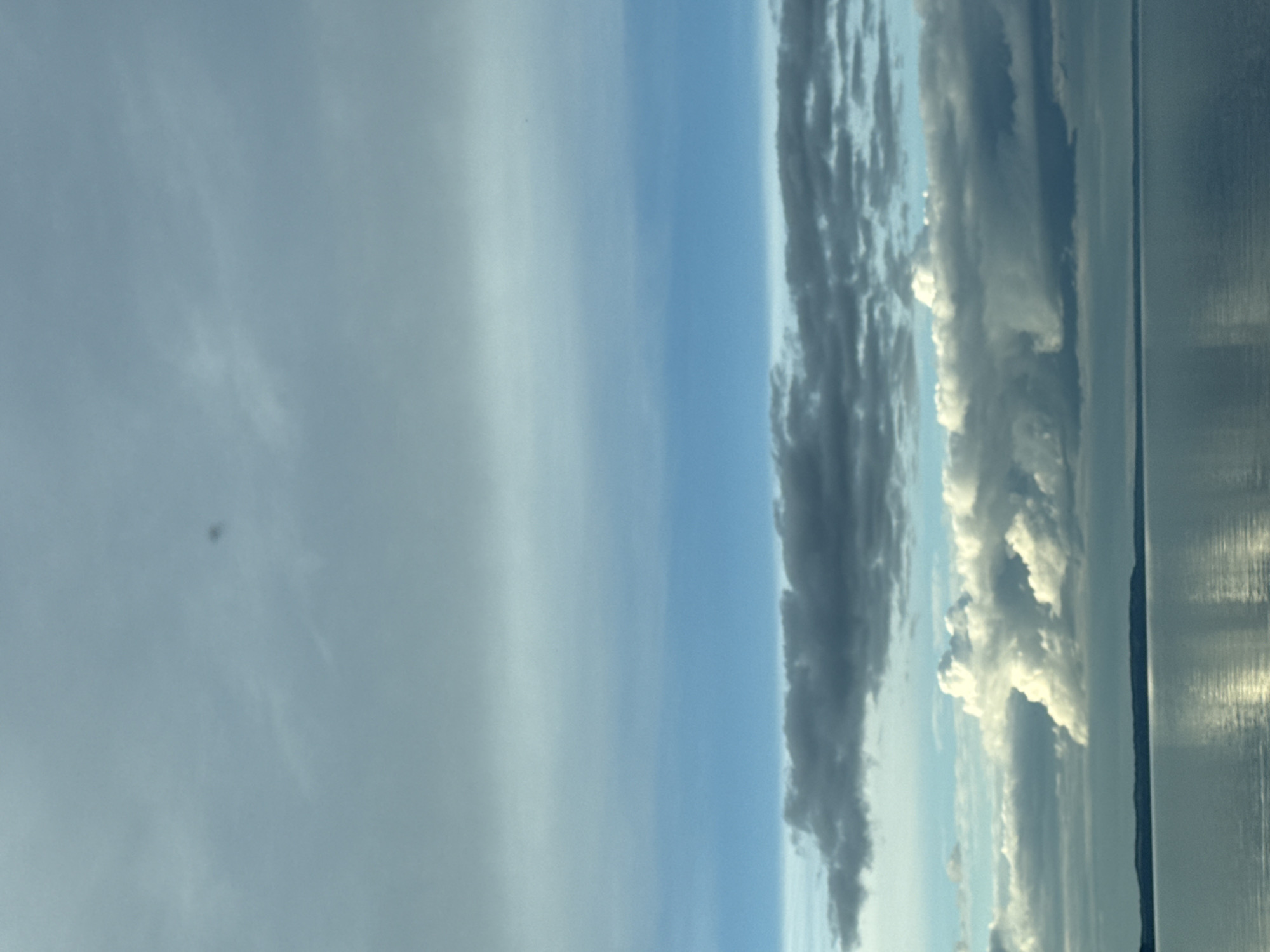 The image shows a view of the sky and water, seemingly taken from indoors or through a window. The sky is light blue and overcast with long, striated clouds stretching horizontally. Some clouds are dark grey, others are bright white, especially toward the right of the image. The water is a reflective pale grey. The location, Chaikovsky, Russia, cannot be confirmed based on visual data.