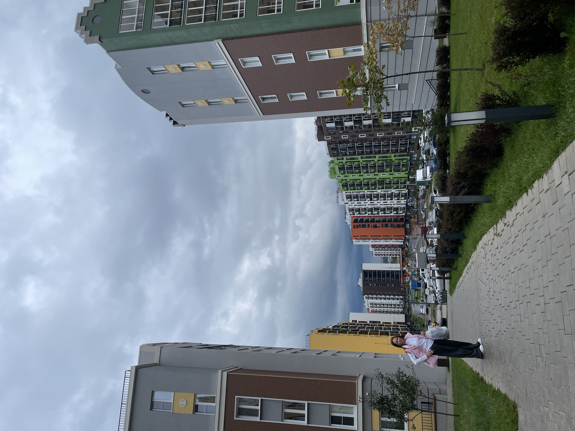 A woman stands on a paved walkway in front of modern, colorful apartment buildings. She is wearing a white long-sleeved shirt, a pink vest, dark pants, and white sneakers. She is carrying a light-colored plastic bag in her left hand. The walkway is made of grey brick pavers and is lined with grass and bushes. Several streetlights are visible along the walkway. In the background, the apartment buildings are tall and multi-colored, with balconies and numerous windows. Cars are parked in front of the buildings. The sky is overcast. The location is the city of Pervomaysky, Russia.