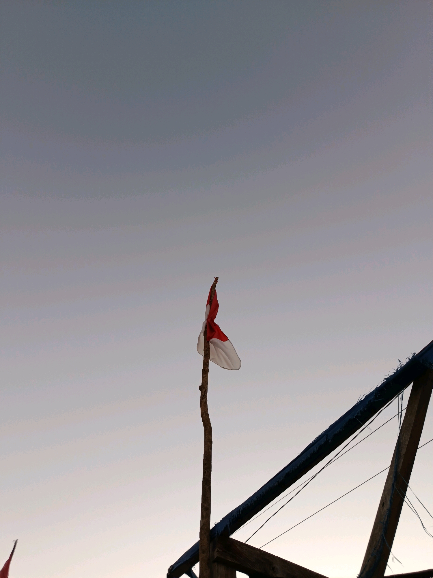 An Indonesian flag, with red on top and white on the bottom, is attached to a rough wooden pole. The flag is partially unfurled and appears weathered. The pole stands against a pale, gradient sky that transitions from a light gray at the top to a lighter, almost white hue near the horizon. To the right of the flag, a section of a wooden structure with dark blue trim and some loose strings is visible, angled upwards and to the right. The scene suggests an outdoor setting, possibly near a coastal area given the context of Bulukumba, Indonesia, known for its shipyards and maritime culture. The overall lighting indicates it may be early morning or late afternoon.