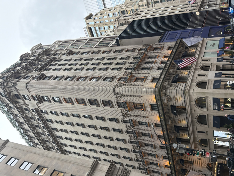 The image depicts The St. Regis New York hotel, located at the intersection of East 55th Street and Fifth Avenue in Midtown Manhattan, New York City, United States of America.
The central subject is the St. Regis building, a multi-story structure with a light-colored stone facade, primarily exhibiting Beaux-Arts architectural styling. Its upper section features a distinctive green, mansard-style roof. The building's facade is highly detailed with numerous rectangular windows, many adorned with small wrought-iron balconies or decorative sills. The lower levels of the building are illuminated by warm orange-yellow uplighting.
At street level, the building houses retail establishments, including a prominent "Harry Winston" store, identified by its signage and dark awnings over arched display windows. An American flag is displayed from a flagpole on the second floor, alongside a plain white flag.
To the immediate left of the St. Regis, a plainer, taller stone building is partially visible. To the right, an adjacent building with a dark, vertically ribbed facade is present, followed by other multi-story buildings of varying architectural styles in the background, under an overcast grey sky.
The foreground includes a street intersection with a traffic light and street signs, indicating "55 ST" and "ONE WAY." Several pedestrians are visible on the sidewalk, some holding umbrellas, suggesting damp or wet conditions. A dark vehicle is partially visible on the street. The perspective is low-angle, looking upwards at the buildings.