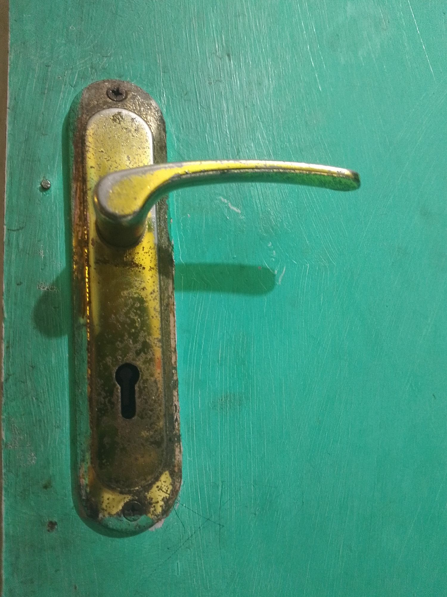 The image displays a close-up view of a tarnished, gold-colored door handle with a keyhole, affixed to a weathered, teal-painted wooden door. The handle is curved and extends horizontally to the right, casting a shadow on the door. The teal paint on the door shows visible brushstrokes and scratches, suggesting age and wear. The area around the door handle is particularly dirty and worn, with rust and grime accumulated on the metal plate. The scene appears to be an exterior shot, likely of a residential or commercial property. Based on the provided context, this image is located in Mukapayung, Indonesia.